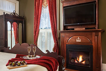 A guest room at the Queen Victoria with champagne and chocolate covered strawberries positioned on a bed and a fireplace beneath a flat-screen TV