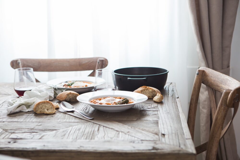 A rustic table with two bowls of stew, chunks of bread, a heavy pot, and two wine glasses