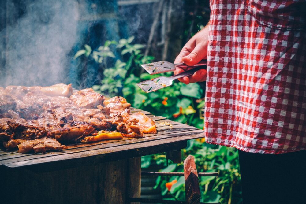 A person in a red and white checkered apron holding tongs and standing in front of meat on a grill