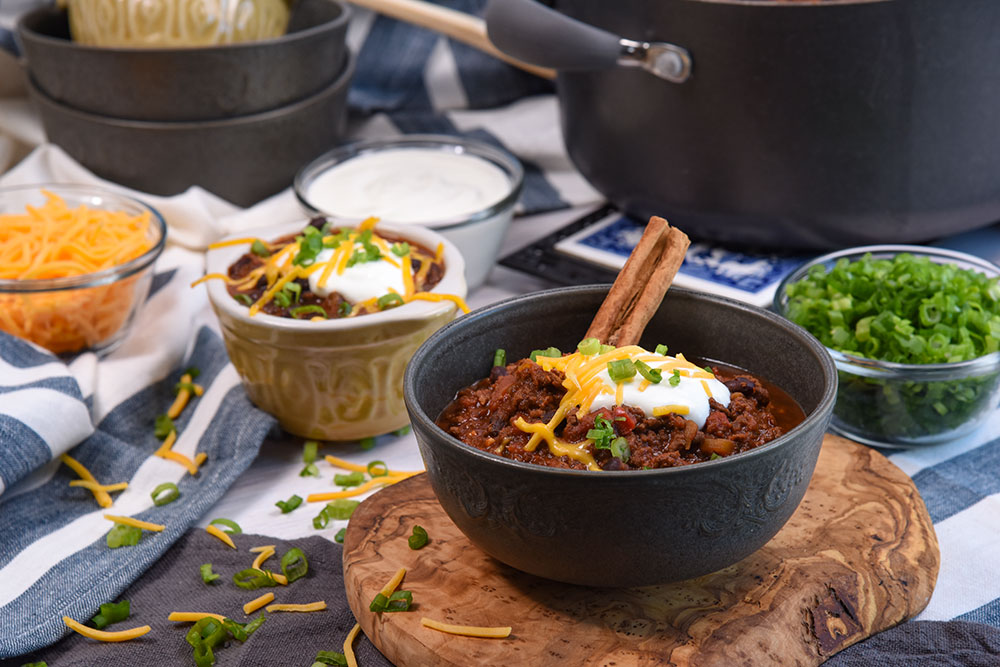 A bowl of chili surrounded by bowls of scallions and cheddar cheese.