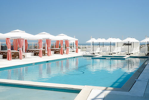 Swimming pool surrounded by whiite and peach cabanas, and white umbrellas and lounge chairs