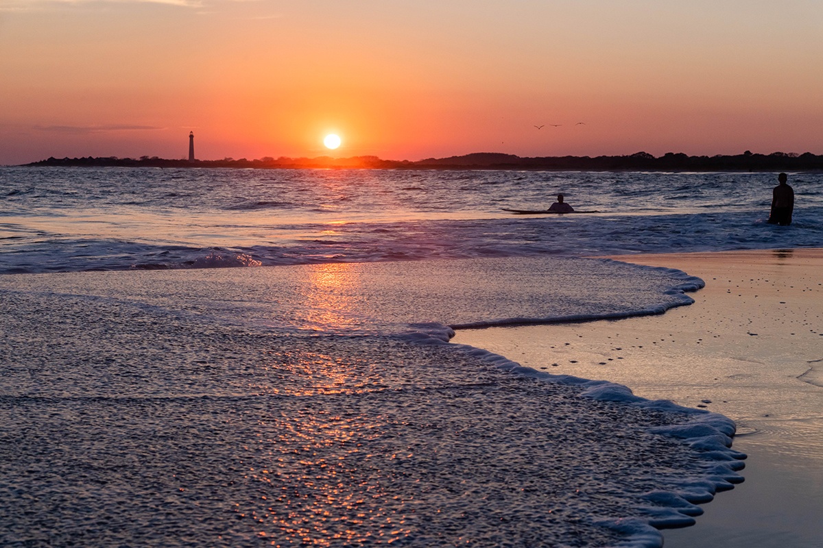 Sunset with a wave rushing in while two people swim and the lighthouse is in the distance