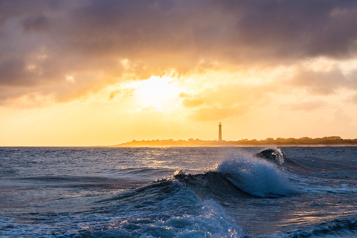 Sunset with waves crashing and clouds in the sky with the Cape May Lighthouse in the distance