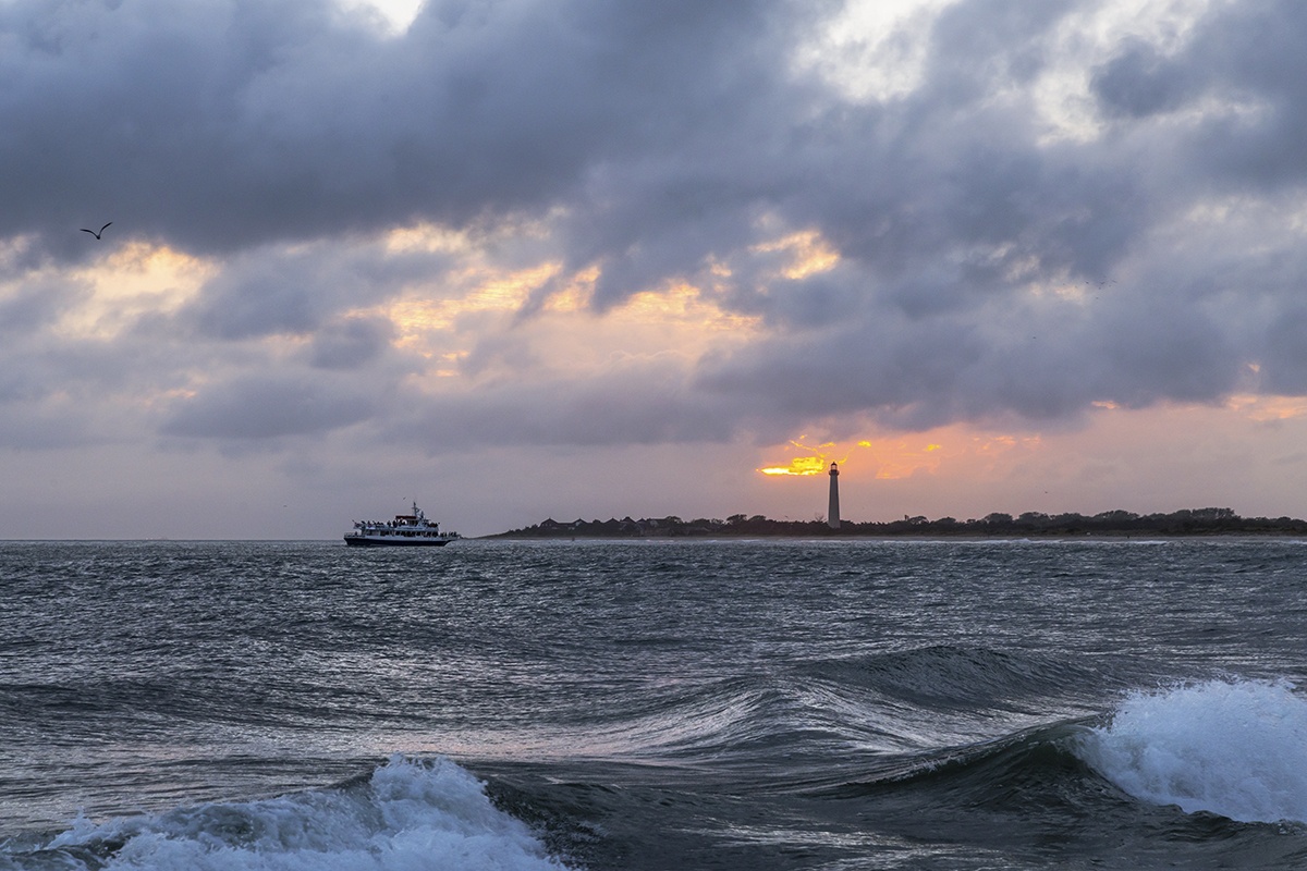 A boat sailing by the lighthouse at sunset with clouds in the sky