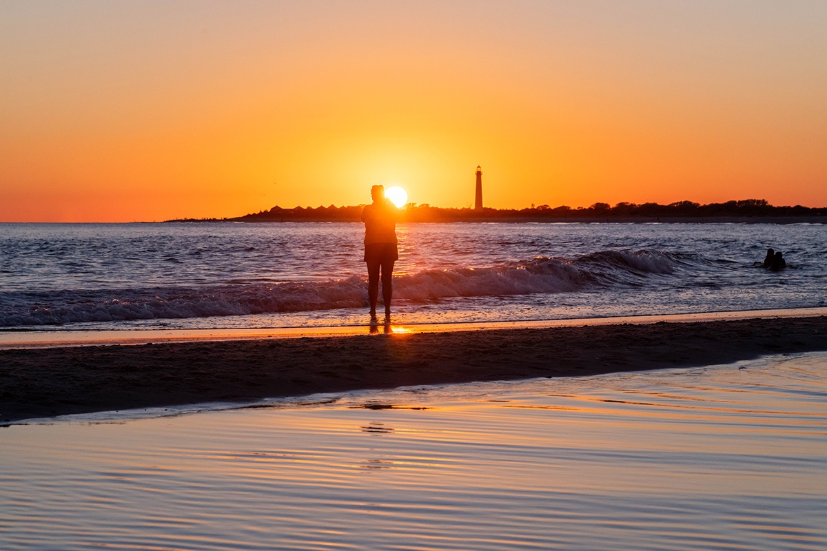 A person silhouetted by the sun setting while waves crash along the shoreline