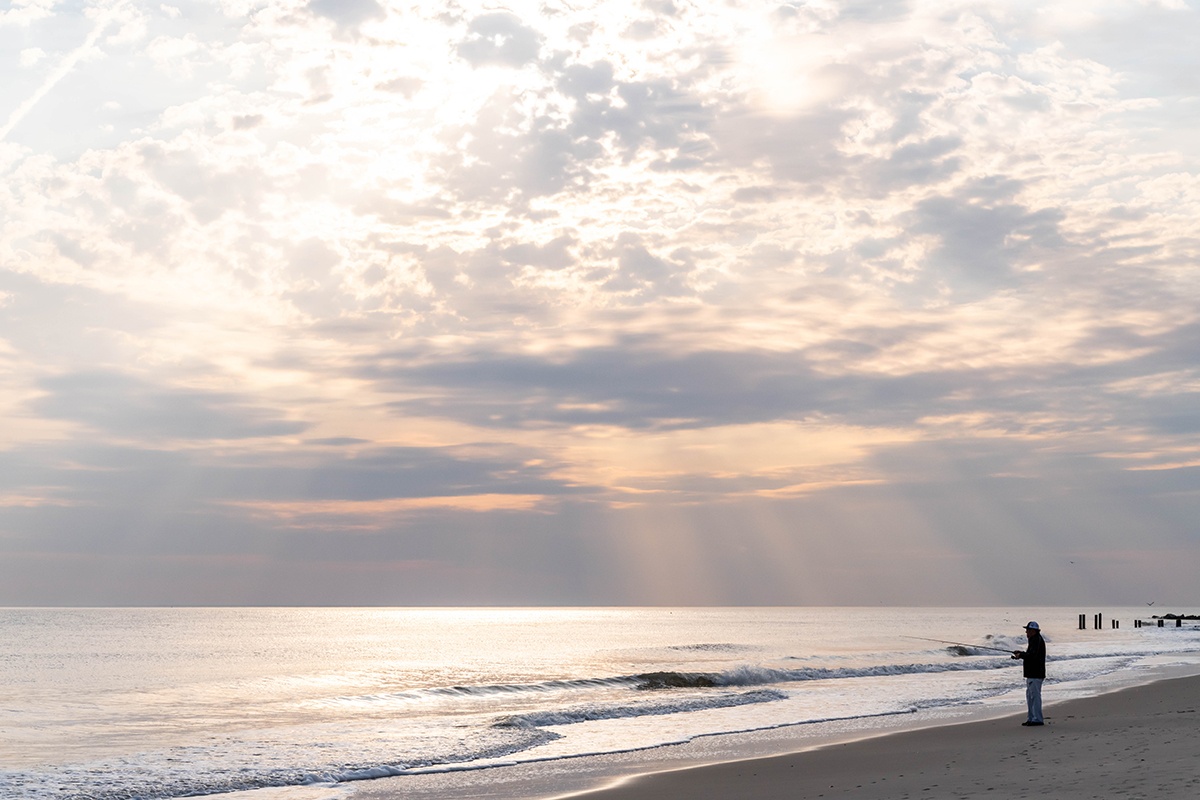 A person fishing at the beach while sun shines through clouds