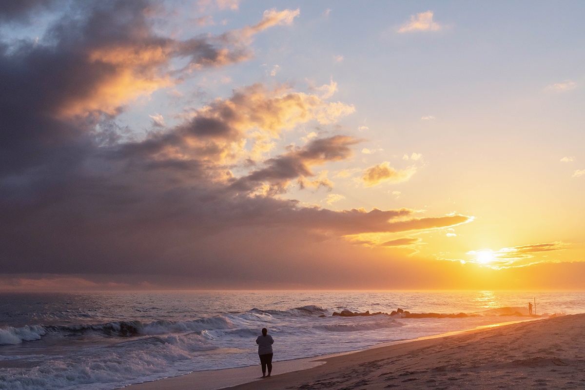 A person walking on the beach as the sun sets with clouds towards the left.