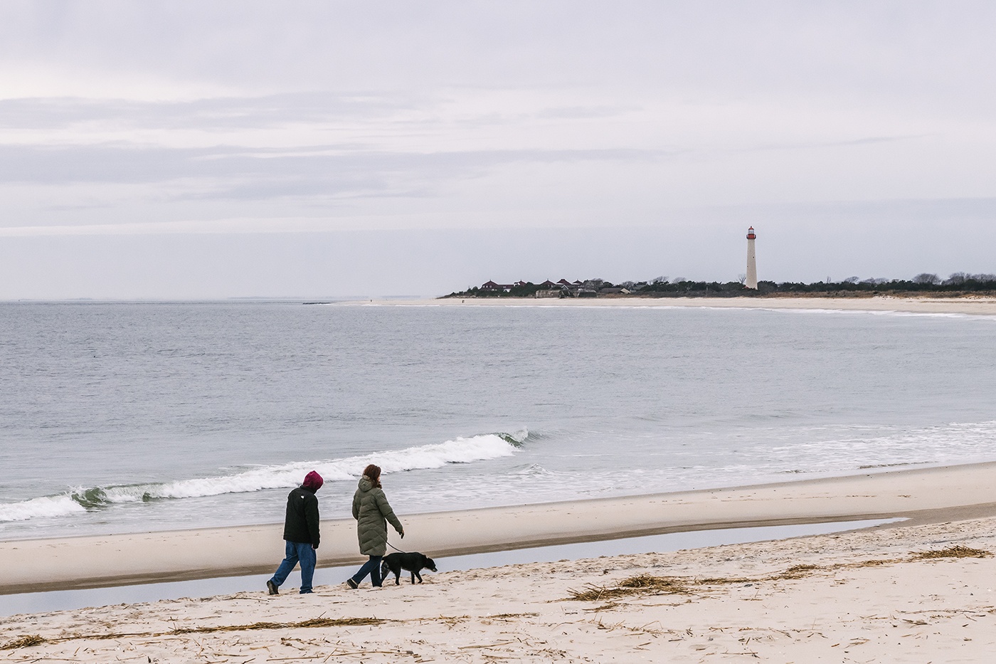 Two people walking with their dog on the beach on a cloudy day with the Cape May Lighthouse in the distance.