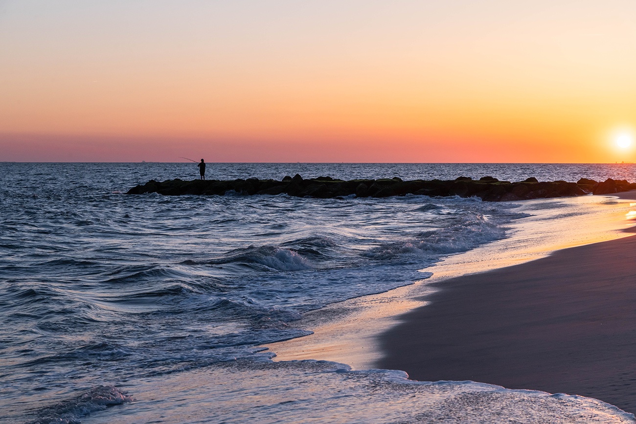 Sun setting while waves crash on the shore and someone fishes in the distance on a jetty