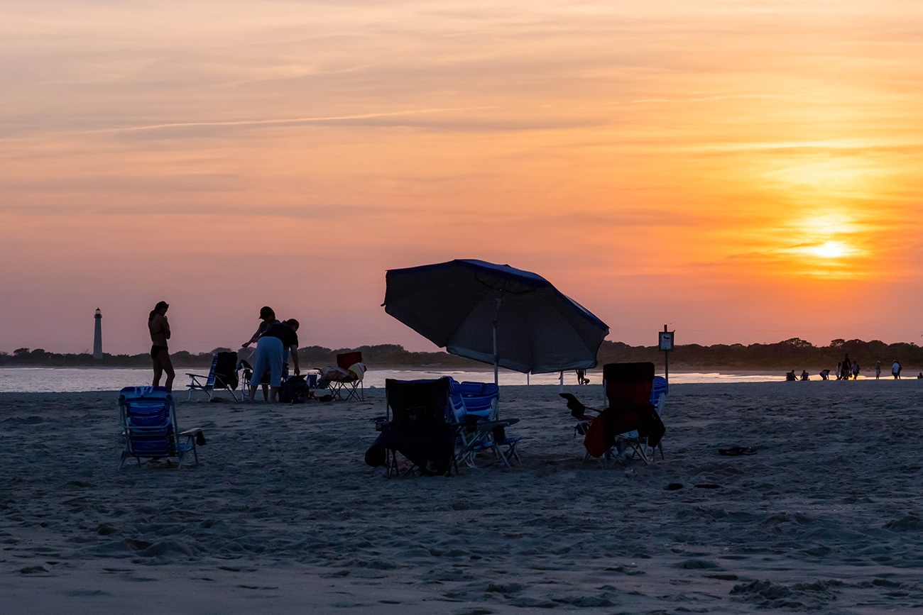 Beach chairs and an umbrella silhouetted by the setting sun with the Cape May lighthouse in the distance