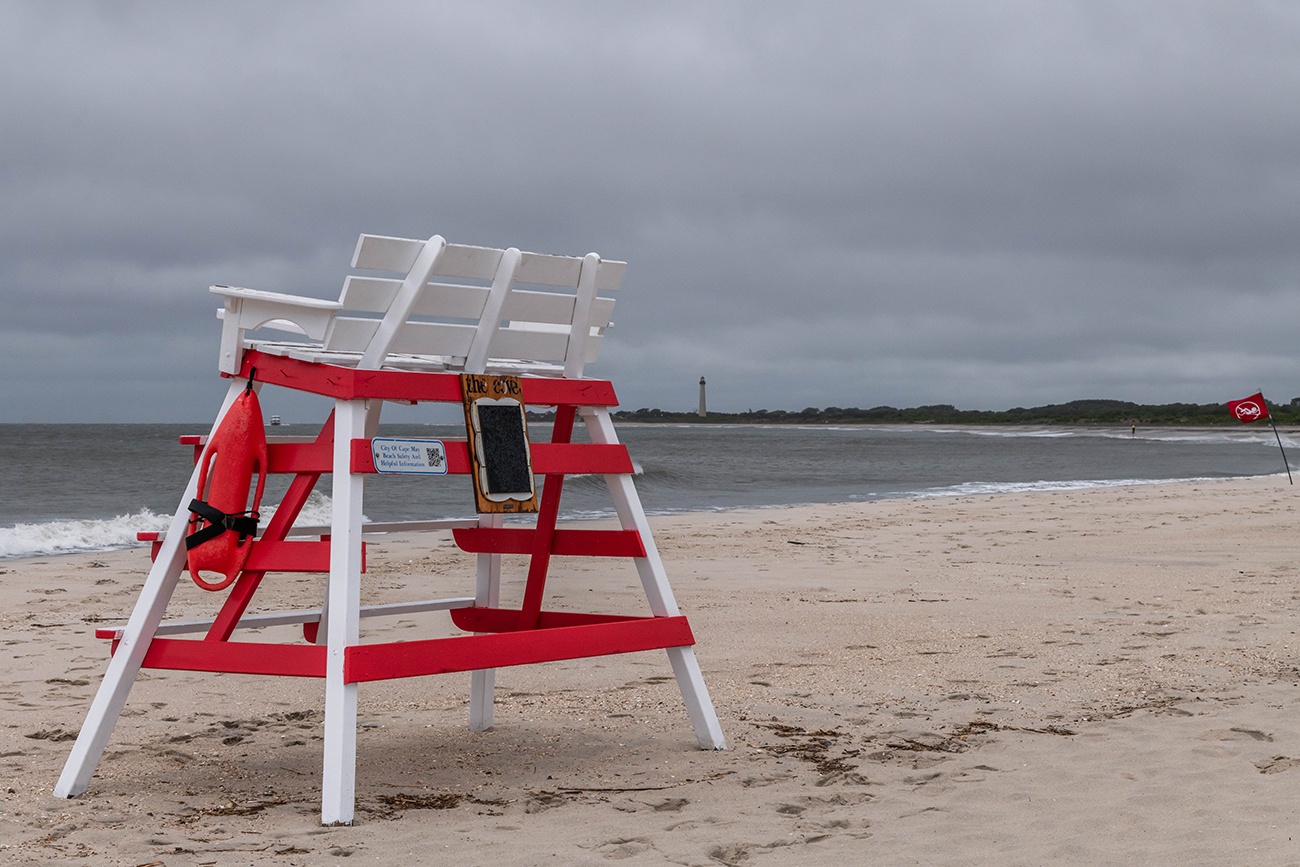 An empty lifeguard stand at The Cove on a rainy and cloudy day with the Cape May Lighthouse in the distance