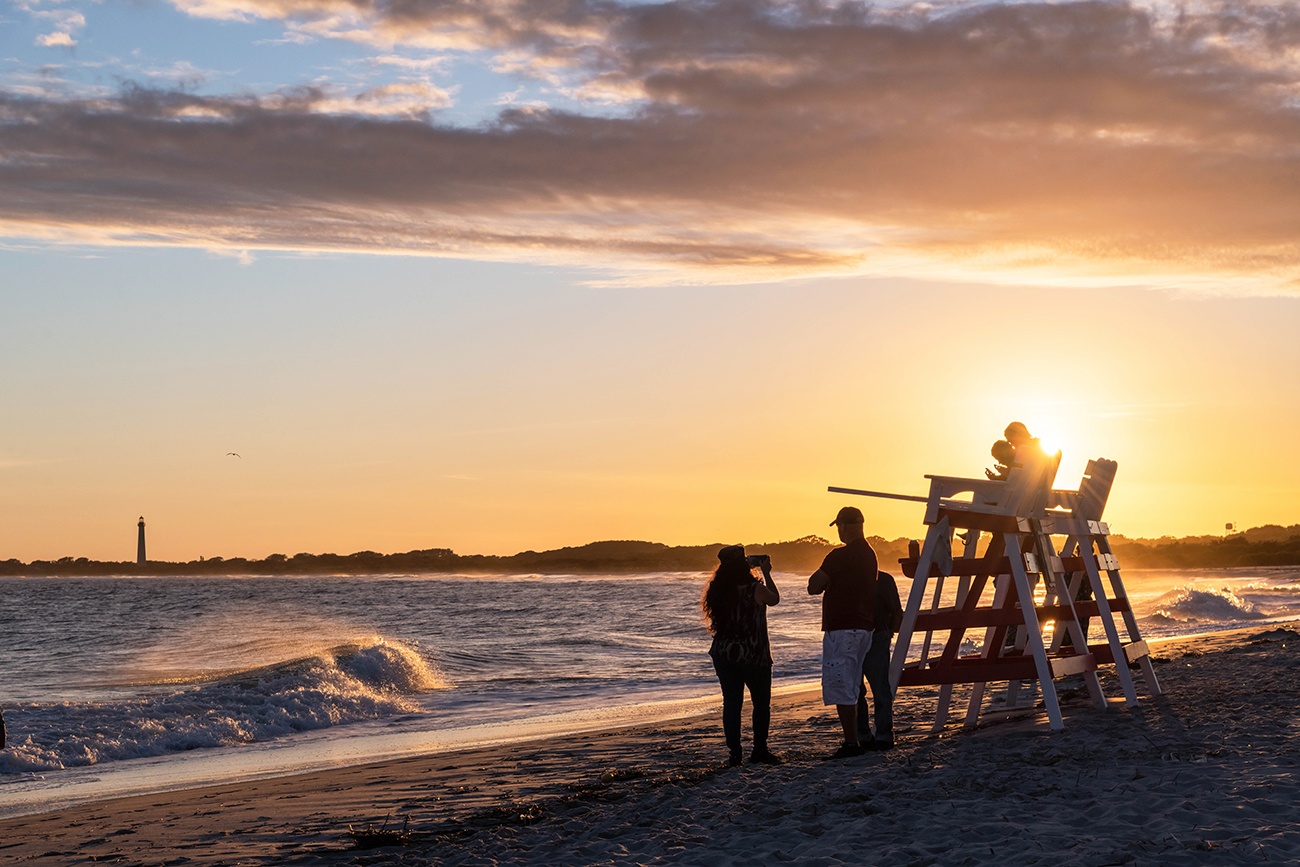 People sitting on lifeguard stands and standing by the ocean watching the sunset as waves crash along the shore