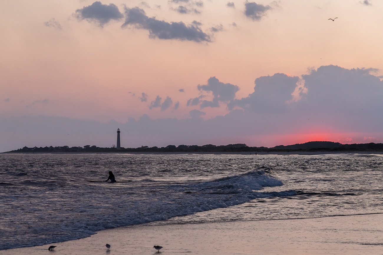 A surfer out in the ocean watching the sun set below the horizon with the Cape May lighthouse in the distance and birds at the shoreline