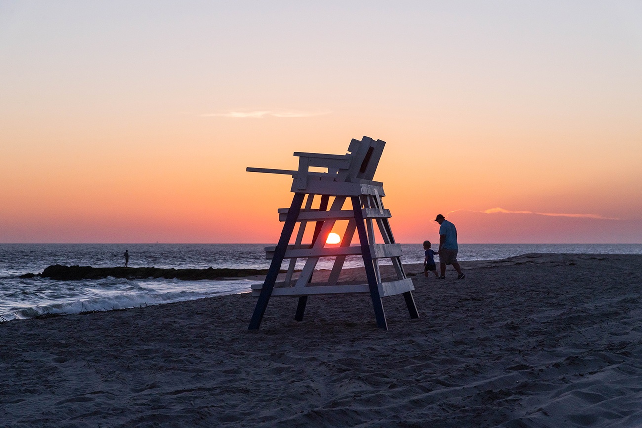 The sun setting behind a lifeguard stand as a father and child walk on the beach