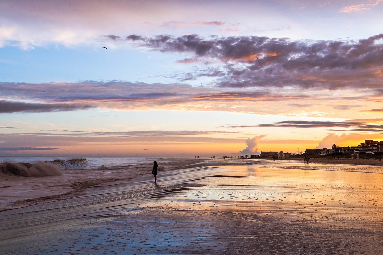 A person standing at the water during high tide as waves crash along the shore at sunset