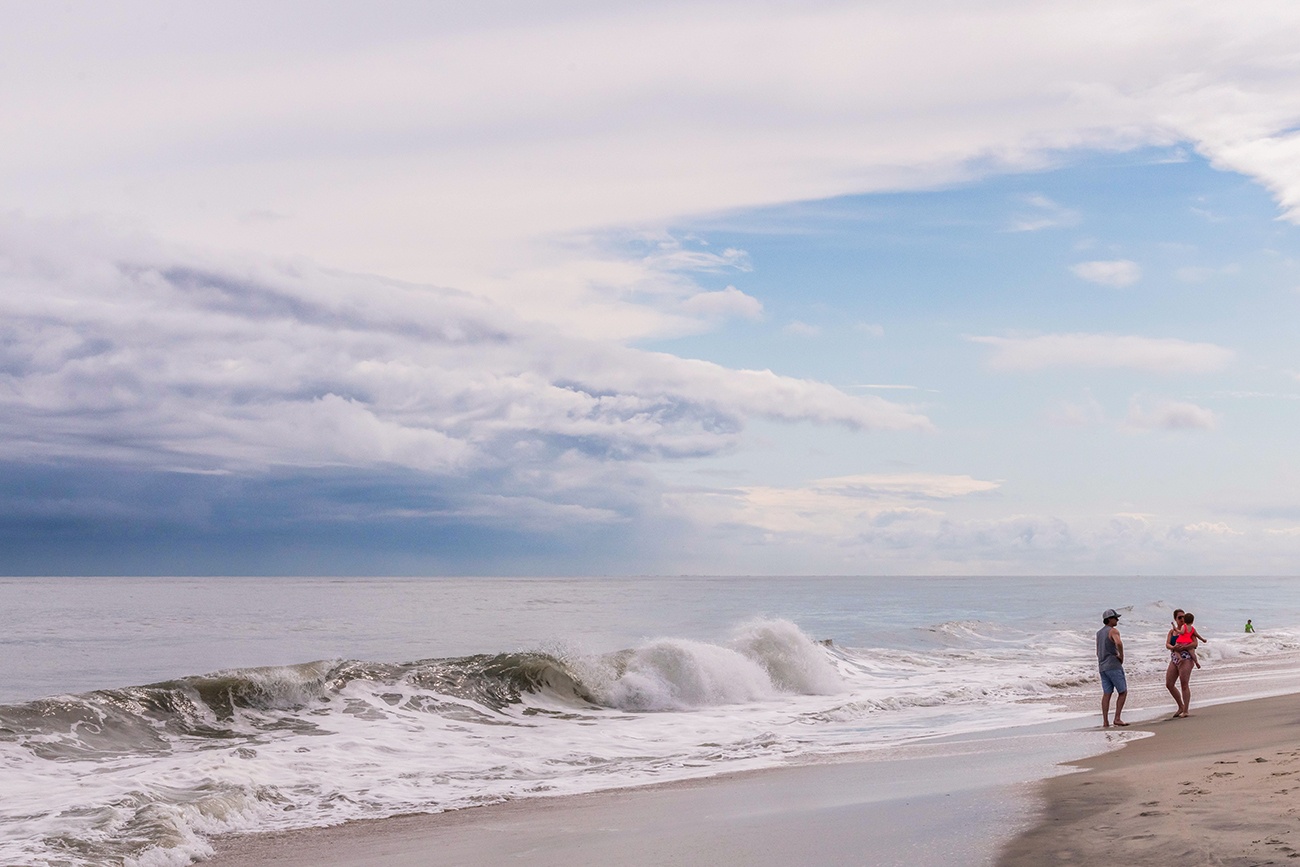 Two people by the ocean as waves crash and dark clouds gather at the horizon