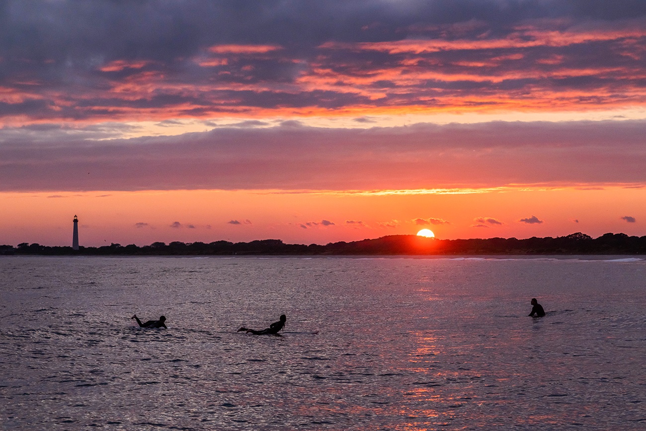 Three surfers in the ocean as the sun sets with the Cape May lighthouse in the distance and pink clouds in the sky