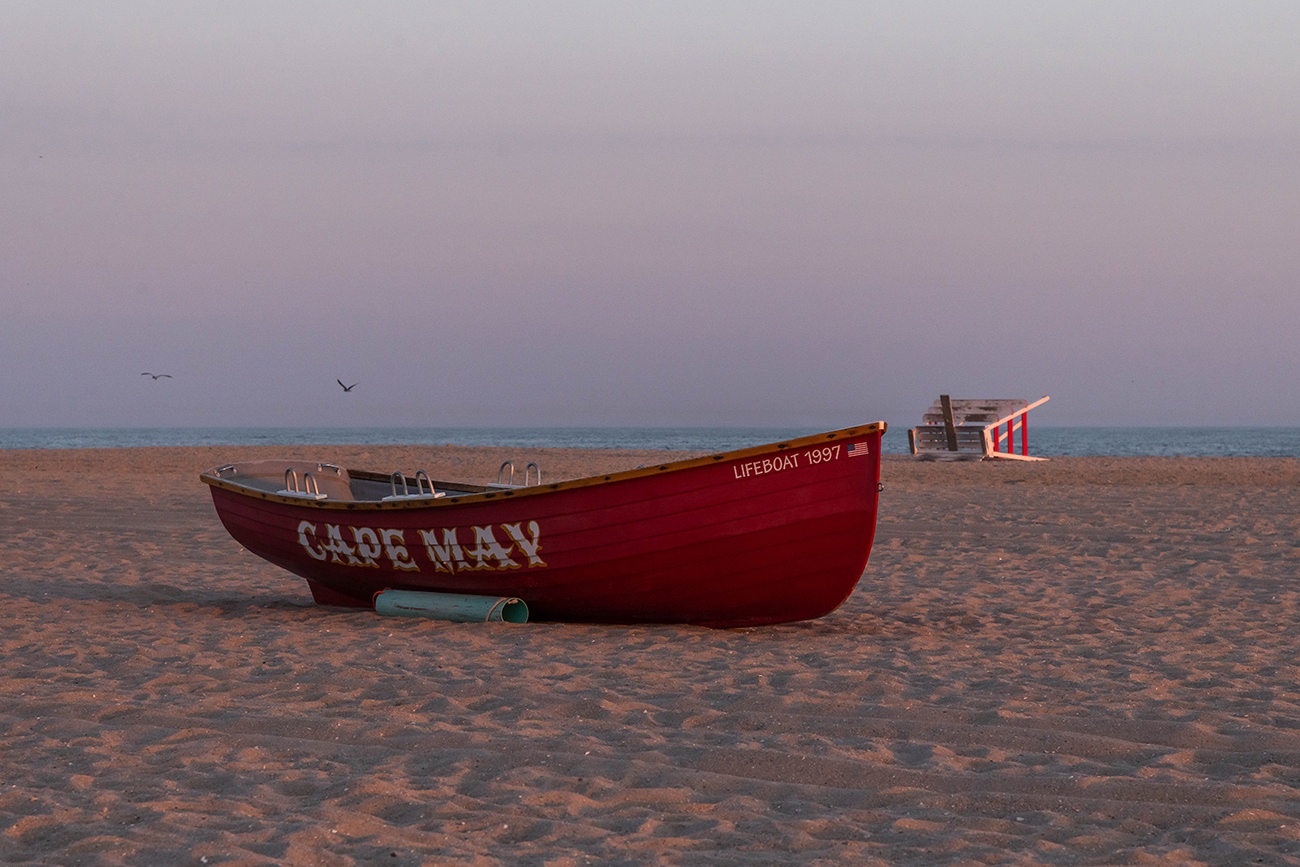 A red Cape May lifeguard boat on the beach with a lifeguard stand down on it's side in the distance at sunset