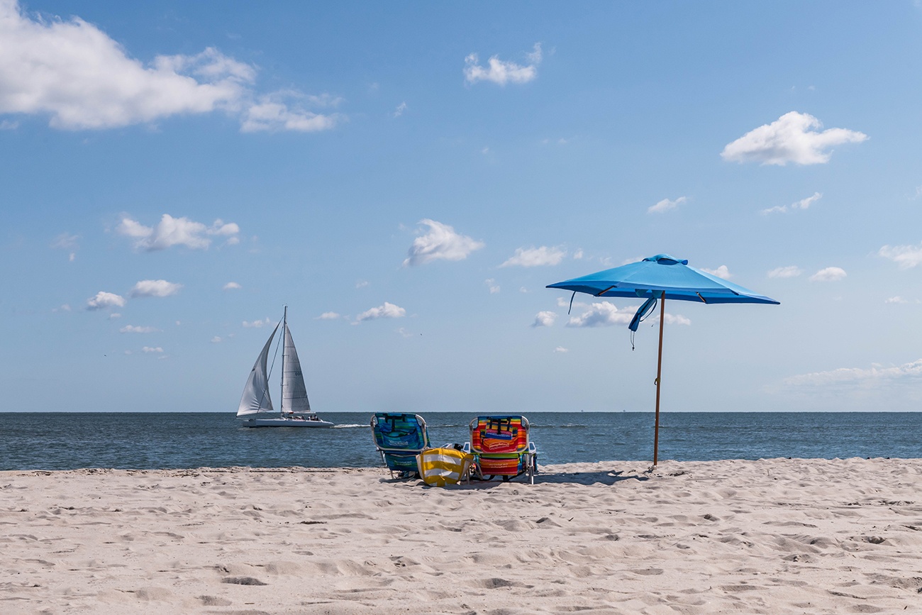 A blue umbrella on the beach with beach chairs and a sail boat on the ocean on a sunny blue sky day