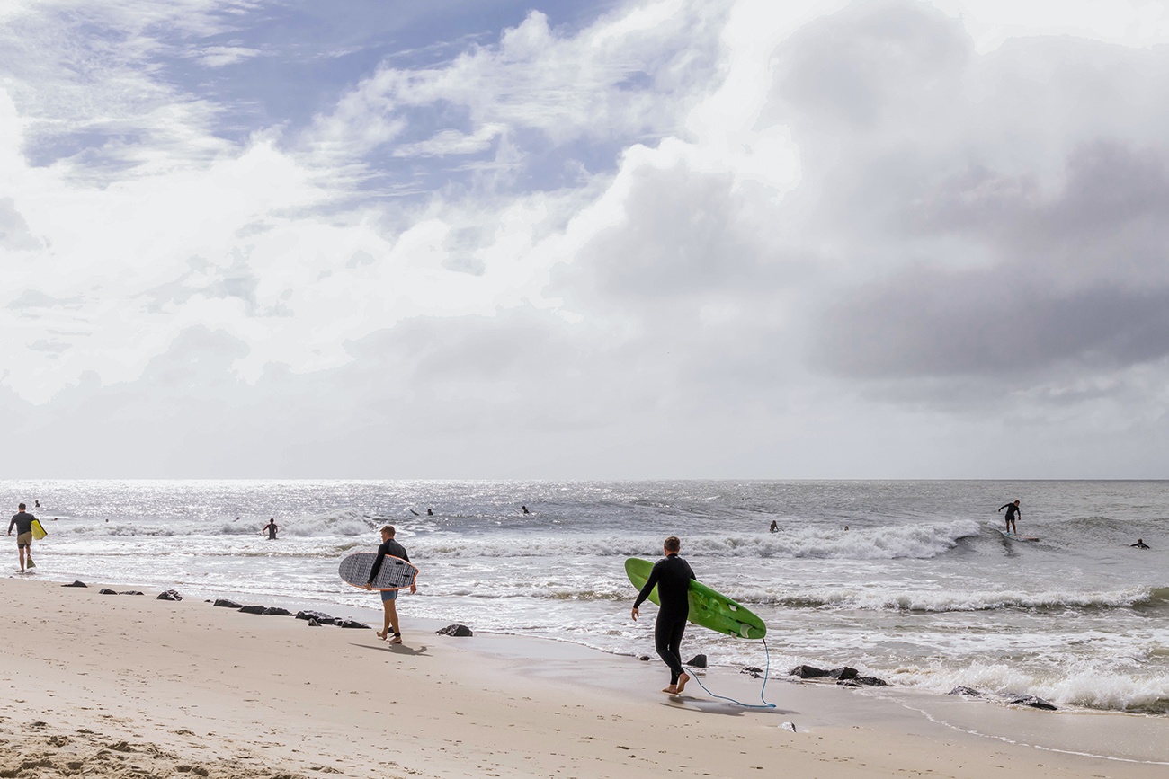 Three surfers walking out to the ocean while other surfers are out in the ocean with clouds and sun in the sky