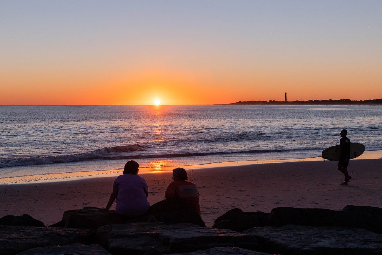 Two people sitting on the jetty watching the sun set as a surfer walks to the ocean with the Cape May Lighthouse in the distance