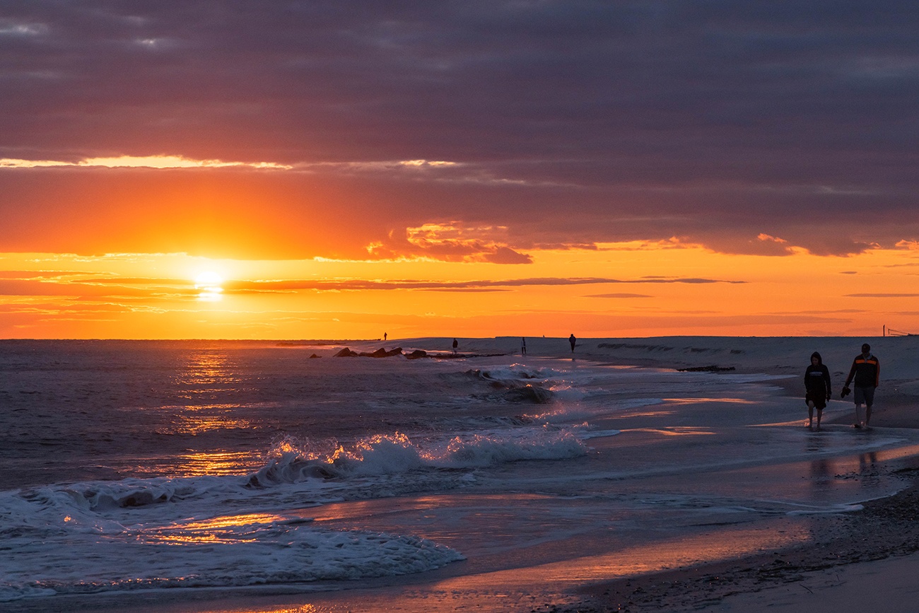 People walking by the ocean as waves crash along the shore and the sun sets with clouds in the sky