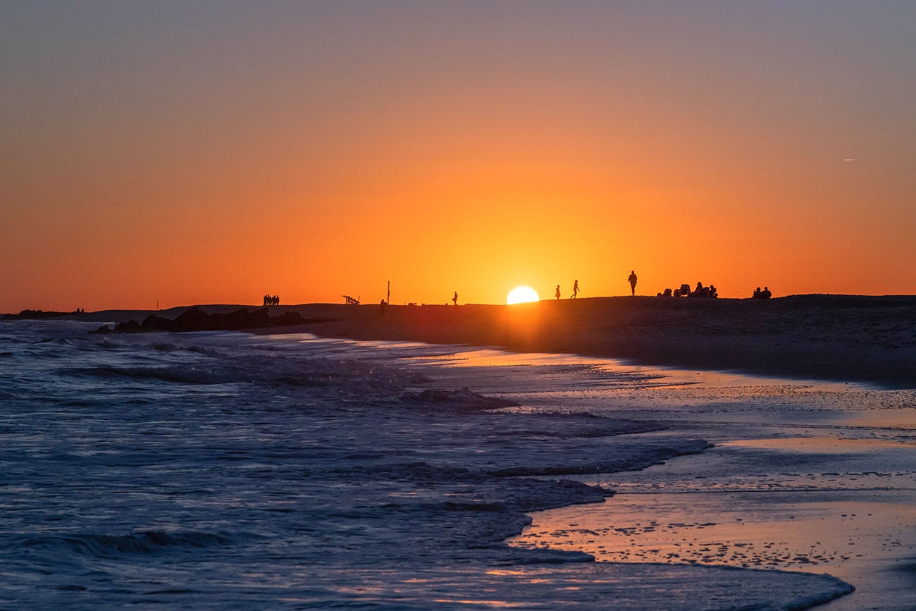 People walking on the beach in the distance with the sun setting behind them with the ocean at the shoreline in the foreground