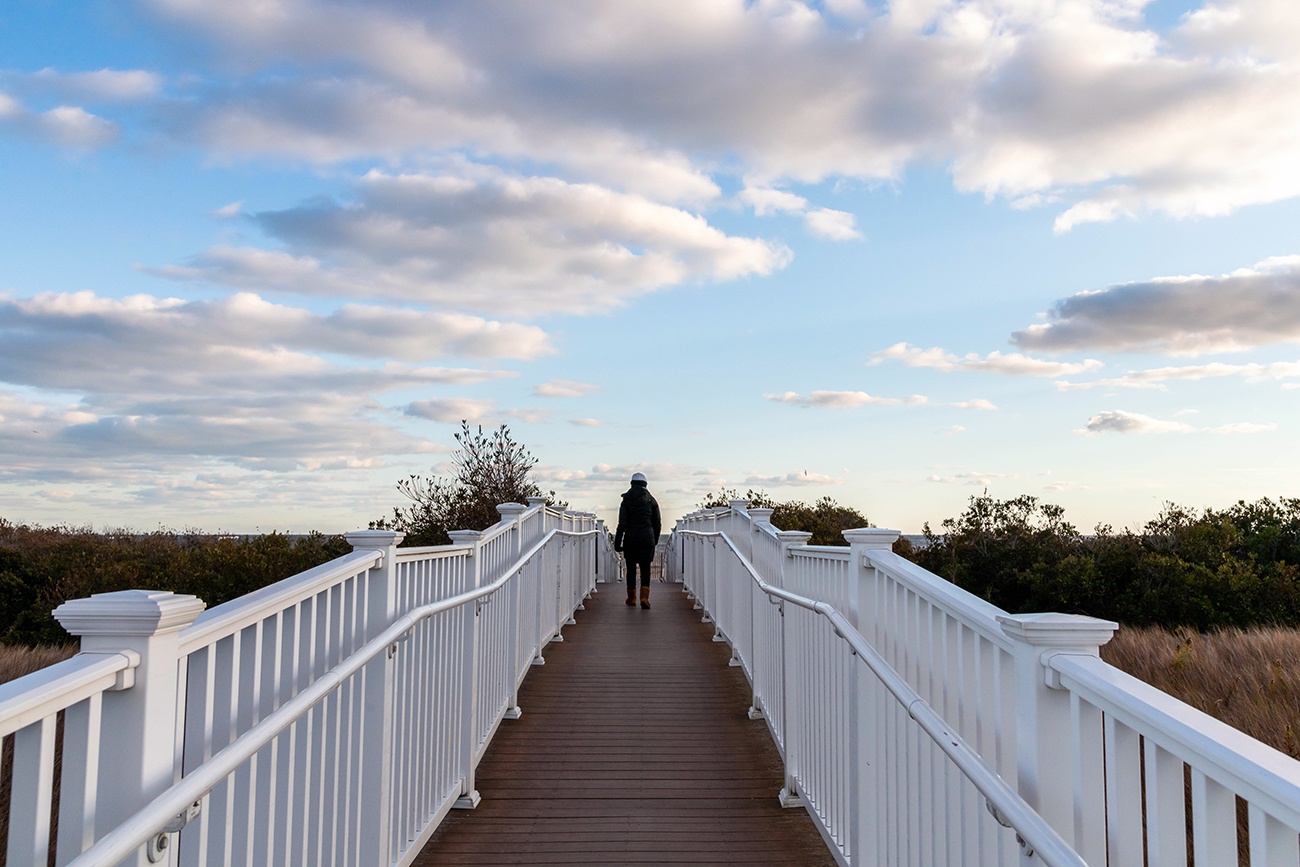 A person wearing a winter jacket, boots, and hat, walking on a path to the beach with puffy clouds in the sky.