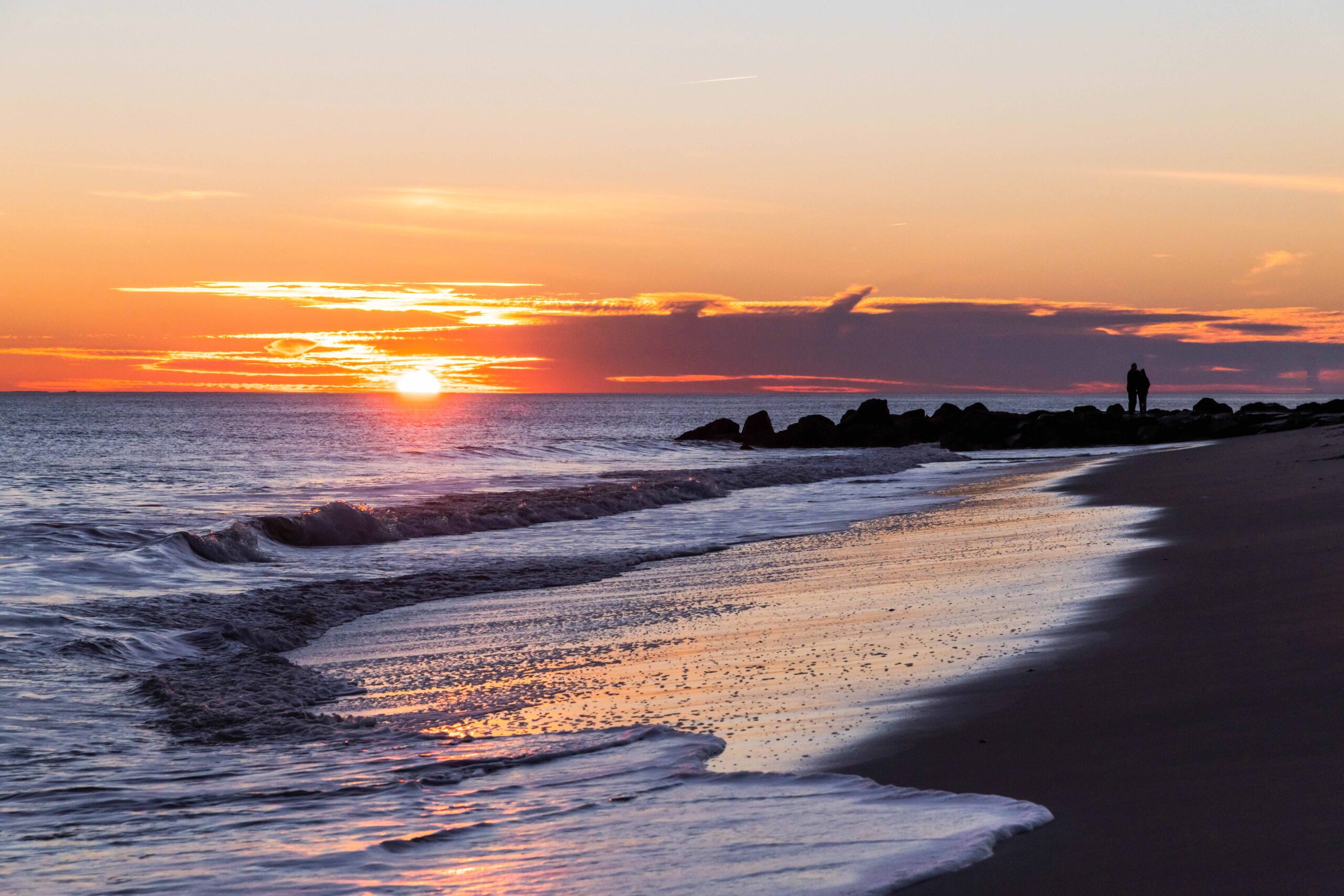 Two people standing on a jetty watching the sun set at the beach while the ocean rushes into the shoreline