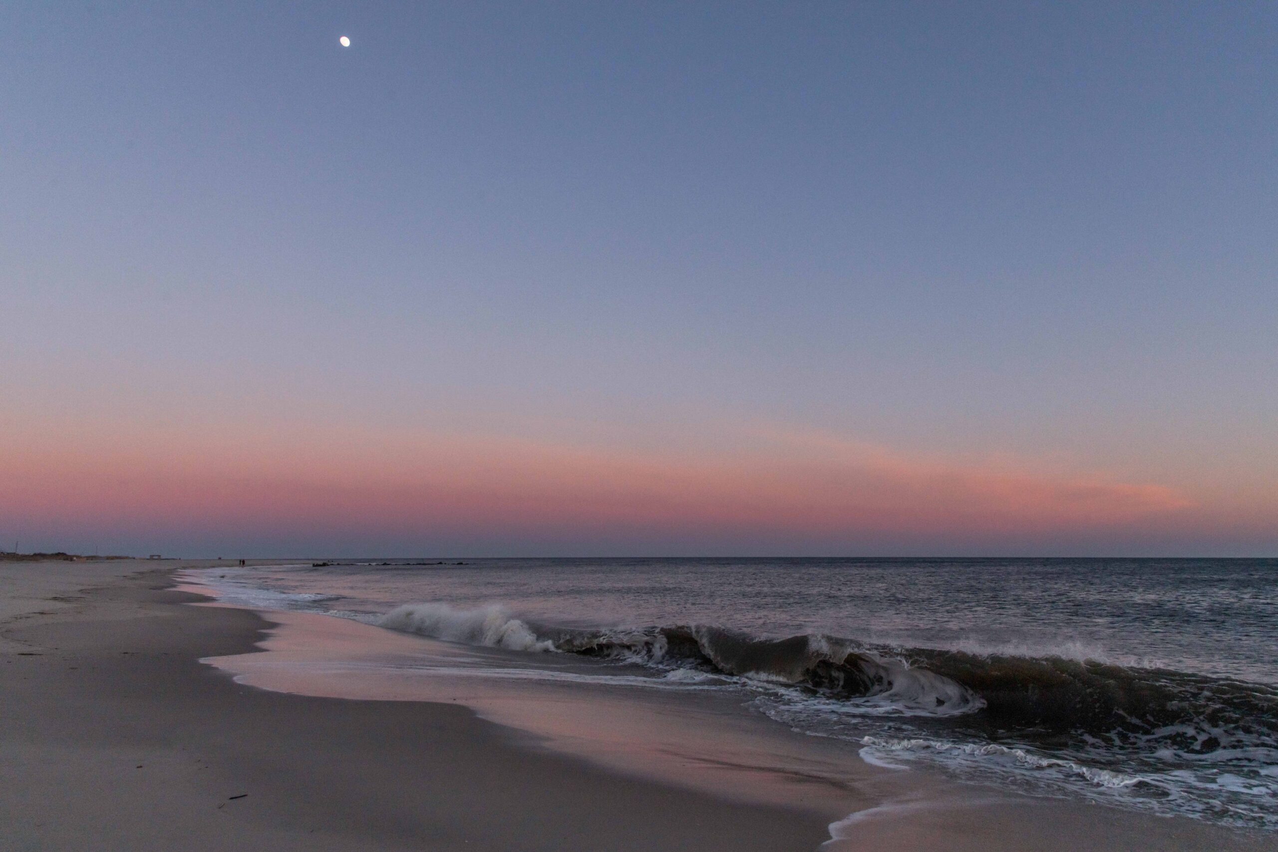 A moon in the sky in a clear sky over the ocean and beach with a wave crashing and pink colors at the horizon