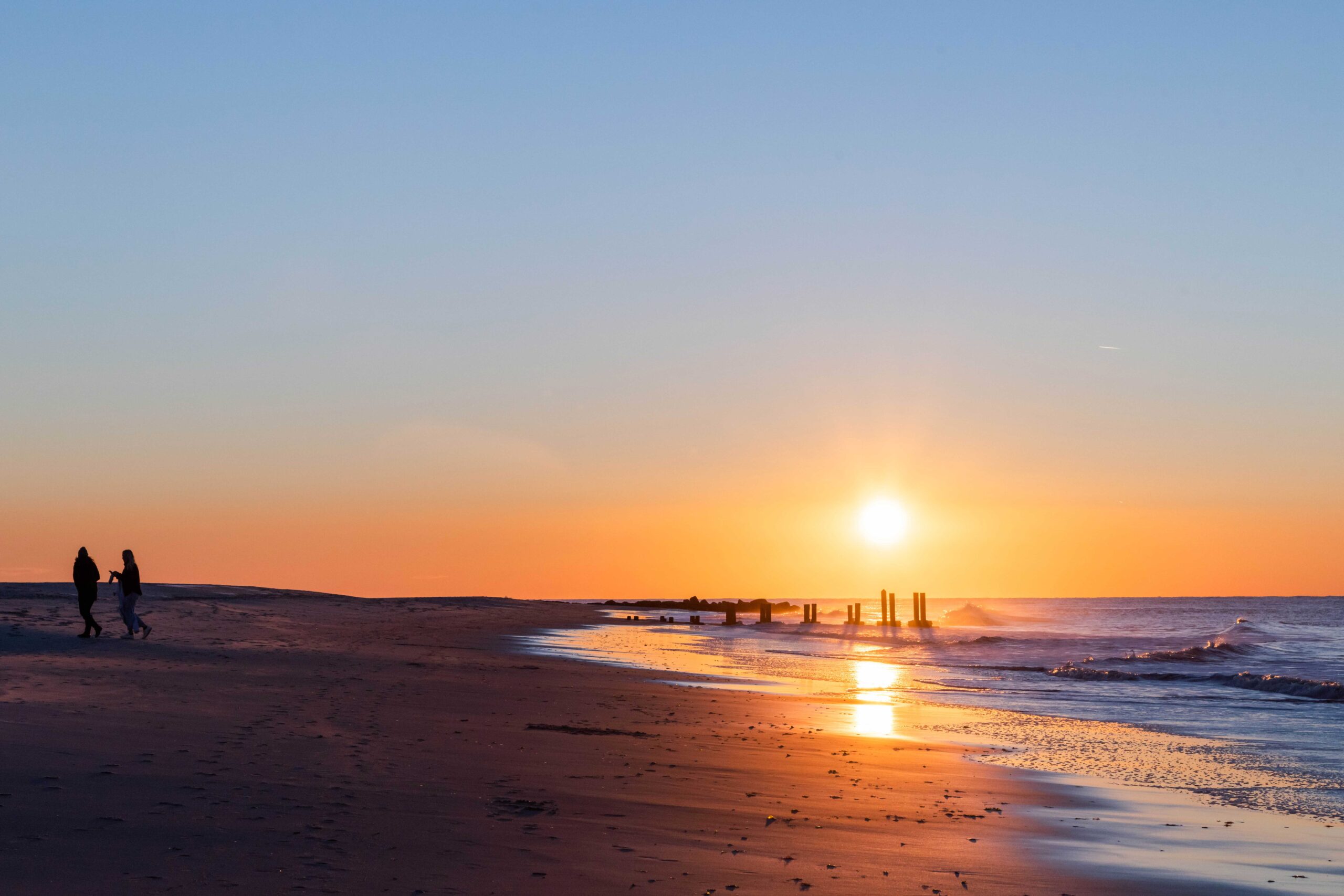 Two people walking on the beach as the sun rises with a clear blue sky and waves rushing into the shoreline