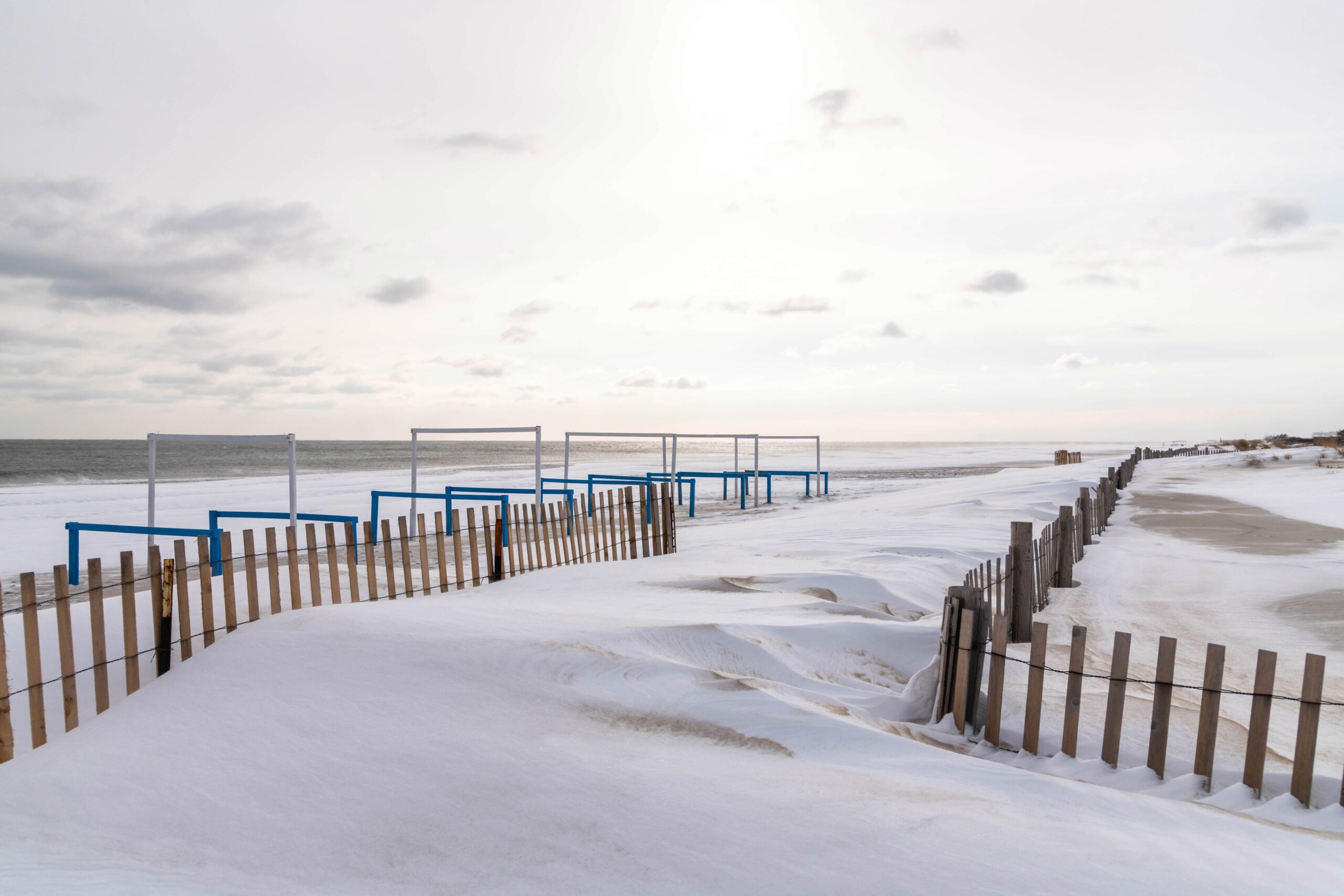 Piles of snow covering wooden fences at the beach entrance with sun streaming through hazy clouds