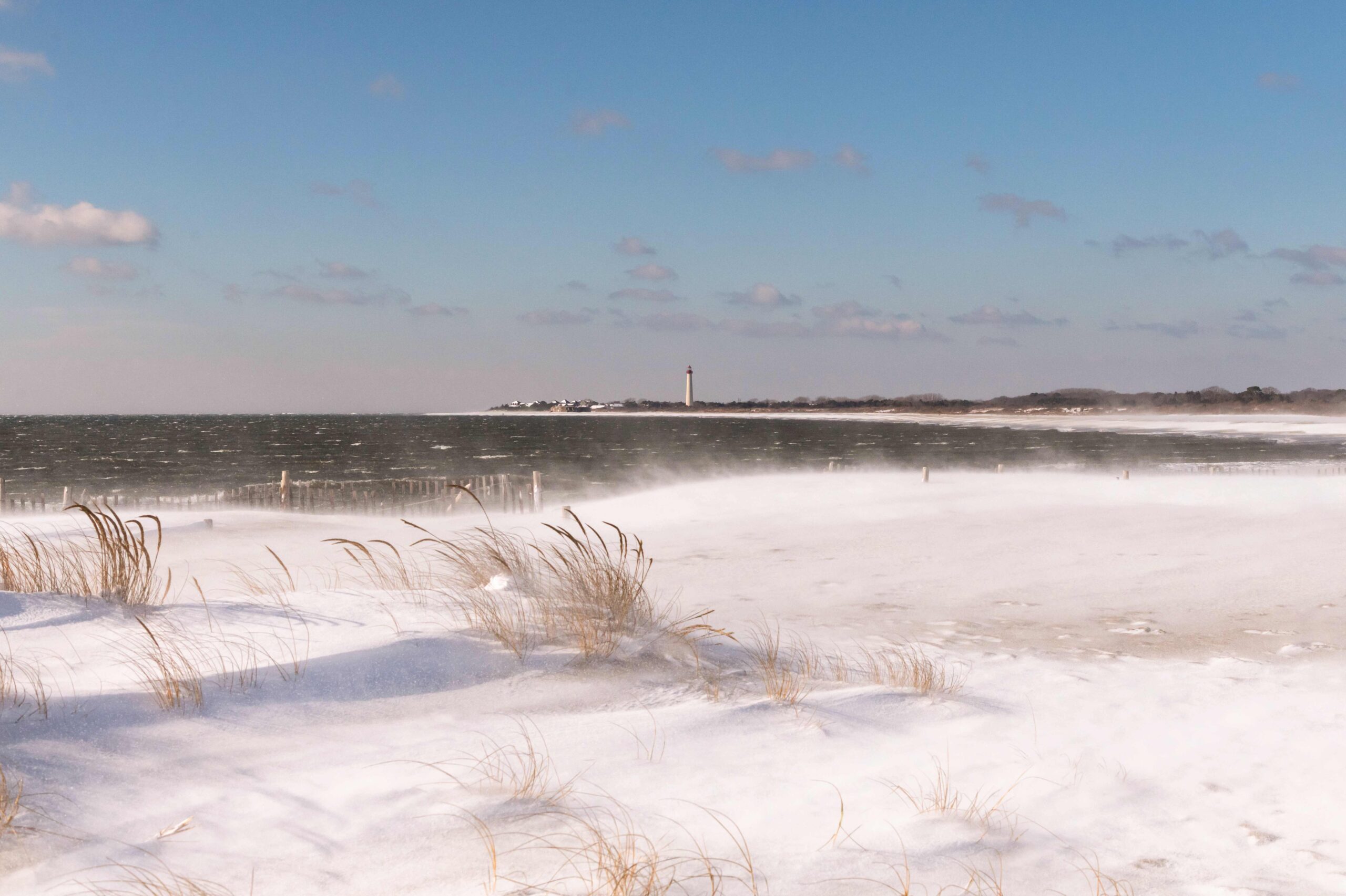 Snow blowing across the sand and dunes with the lighthouse and the choppy ocean in the background