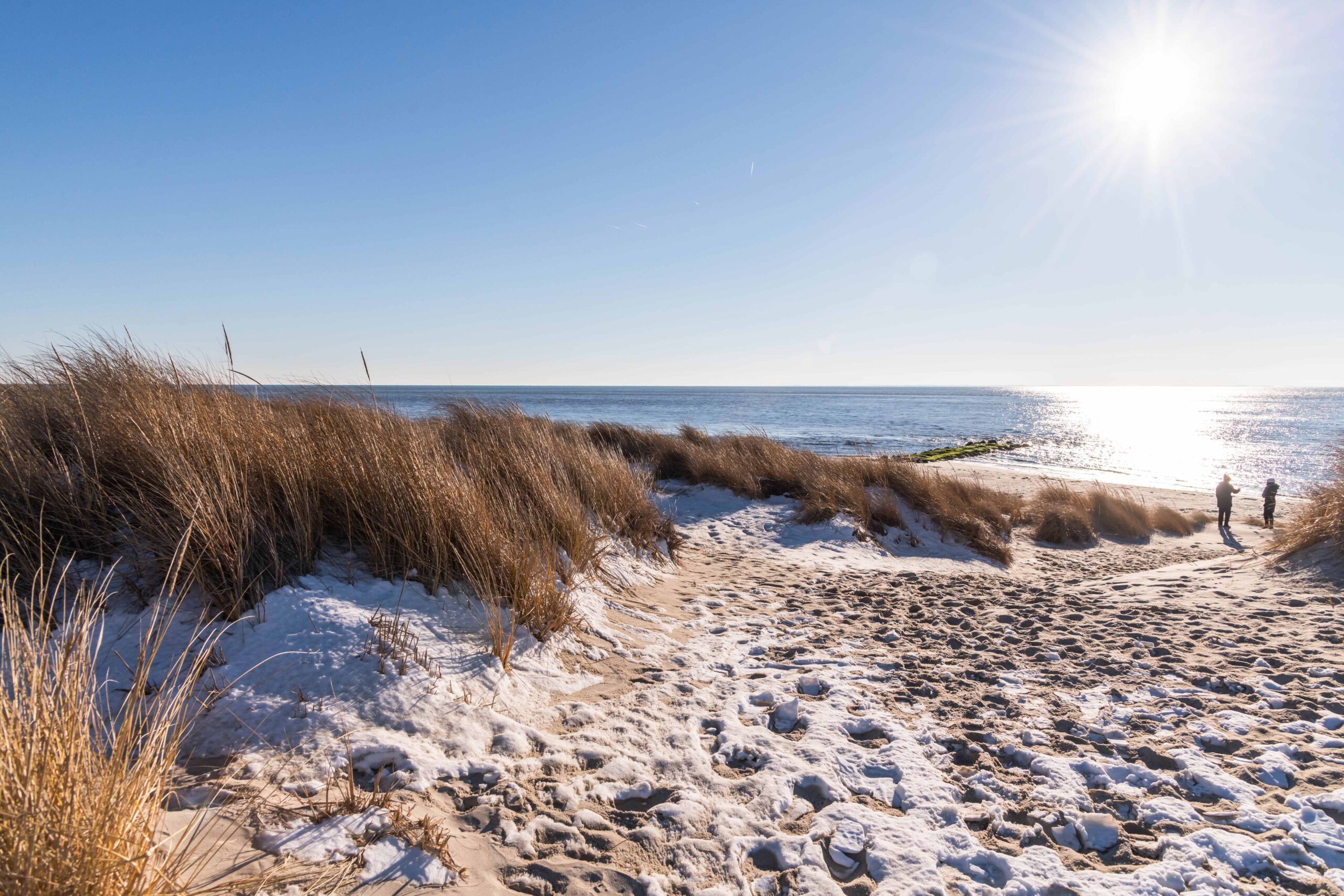 Snow on the sand and in the dunes at the beach with the ocean and two people in the distance on a sunny day with a clear blue sky