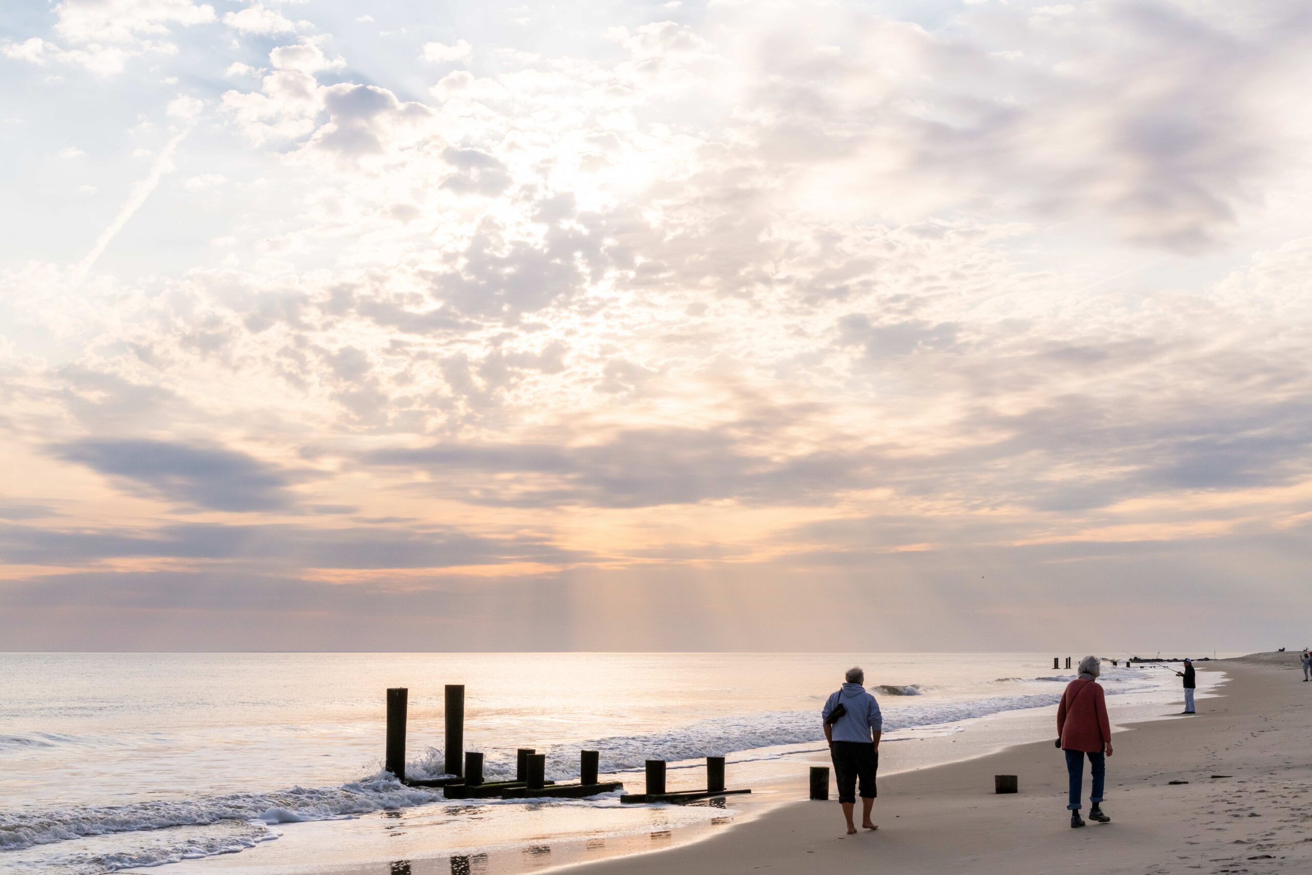 Two people walking on the beach along the ocean with sunlight filtering through clouds in the sky and someone fishing in the distance