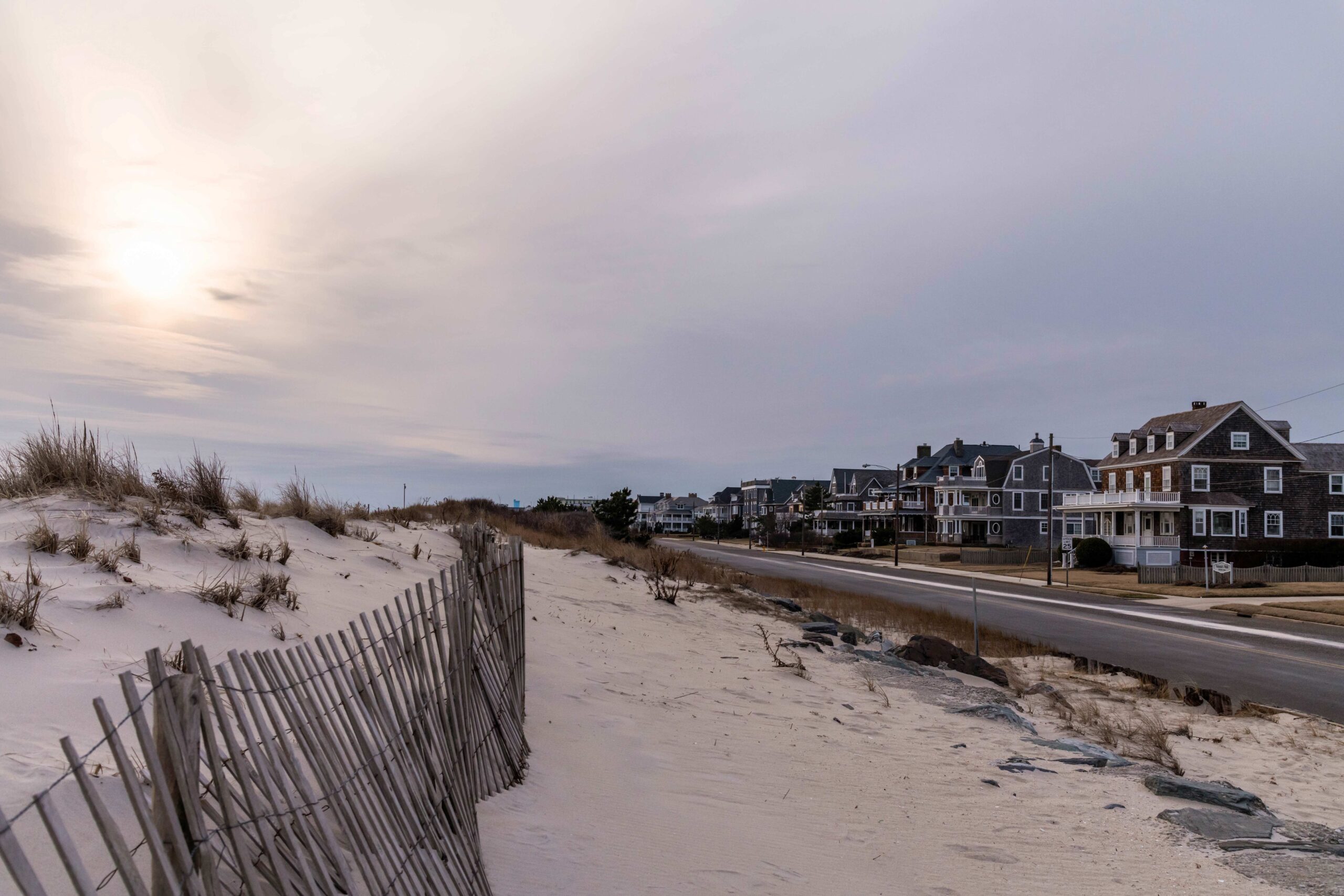 A view down Beach Ave. with cedar shake beach homes on the right and beach dunes with a fence on the left and sun hazily shining behind an overcast blue grey cloudy sky