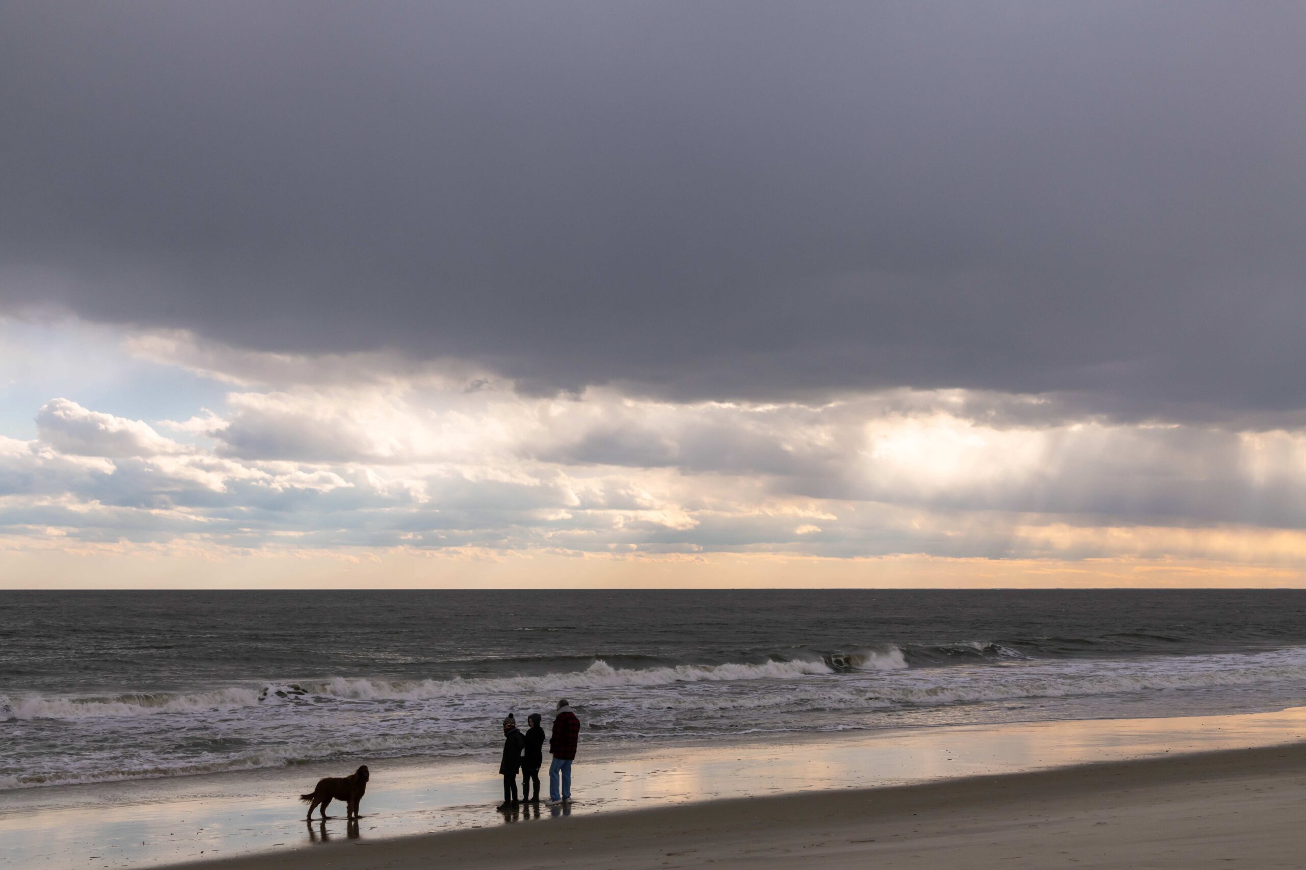 Sunlight streaming through layers of dark clouds in the sky with a family of three and their dog standing at the shoreline looking out at the ocean and sky