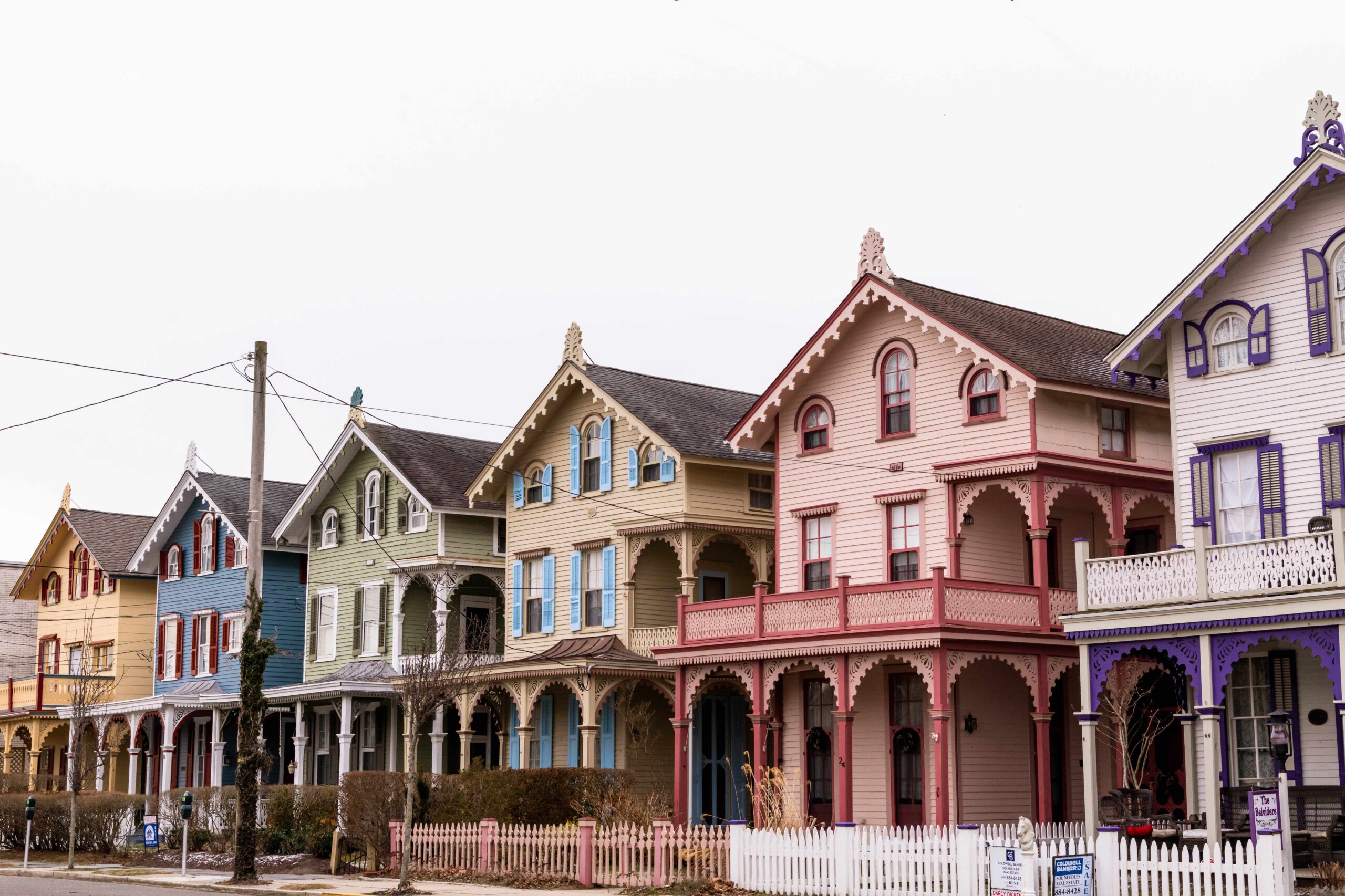 Six colorful Victorian houses in a row (from left to right: yellow, blue, green, tan, pink, purple) on a cloudy day with a gray sky