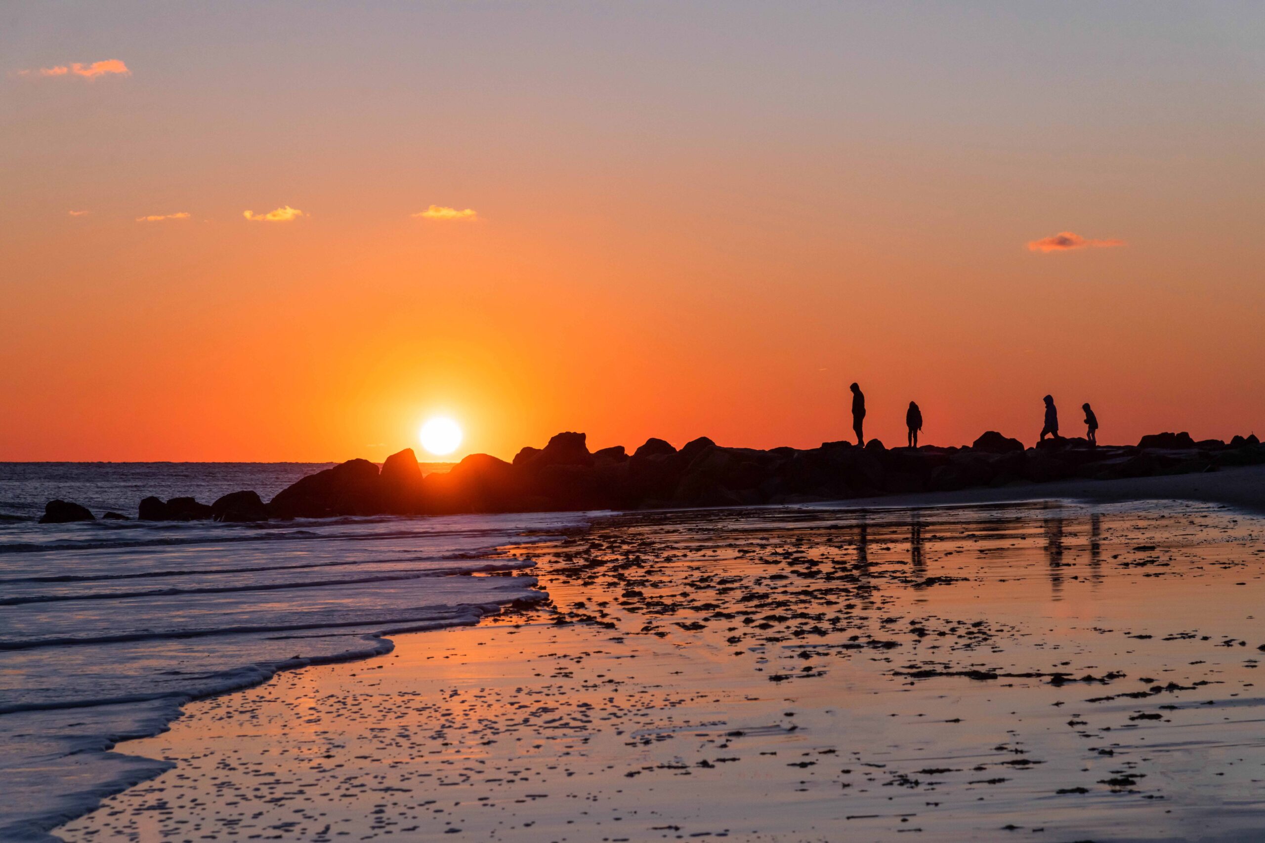 A family of four people walking out on the rock jetty at the beach as the sun sets behind them and the ocean rolls into the shoreline