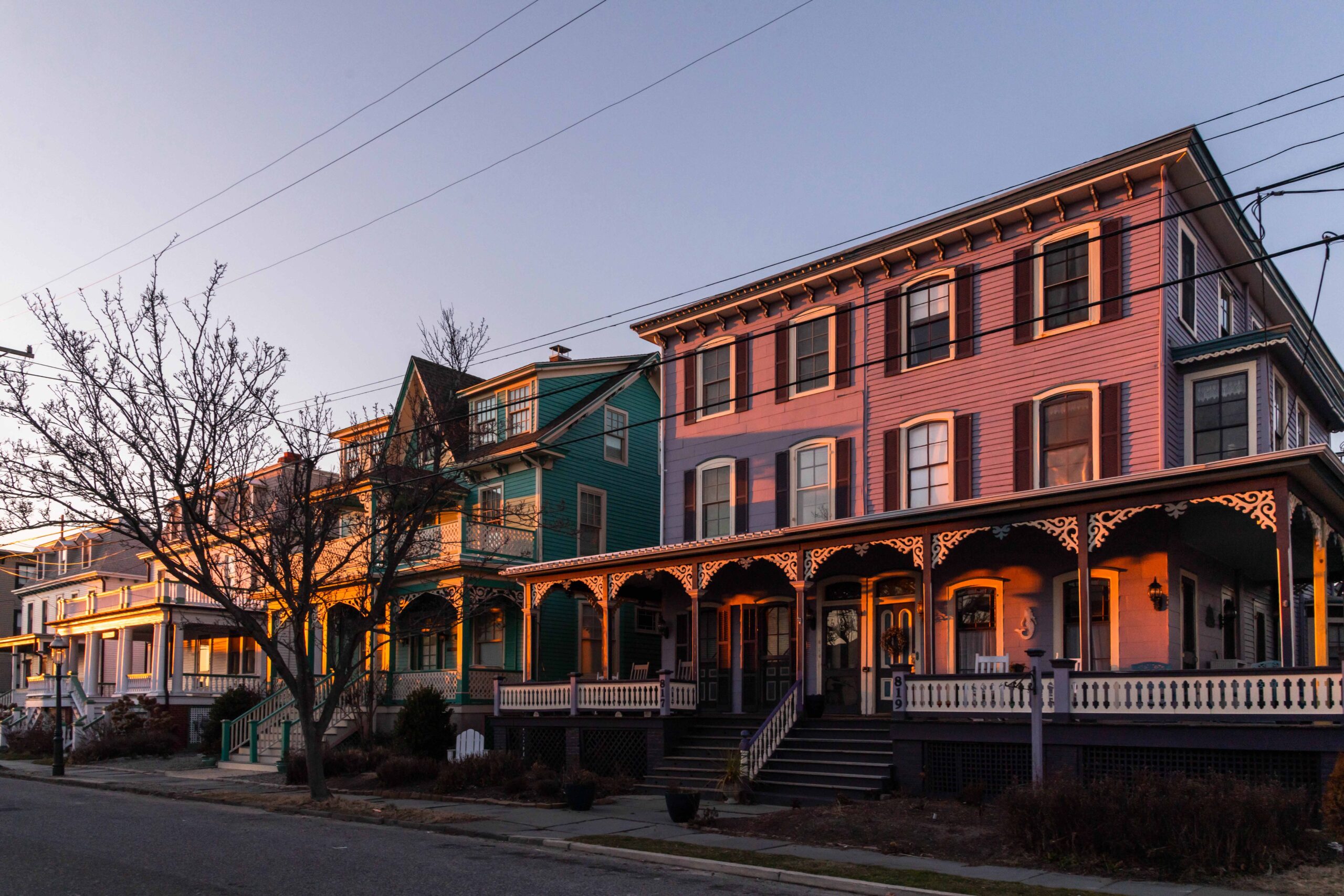 Pink and orange sunset light shining on (from right to left) purple, teal, and white Victorian houses with a clear blue sky