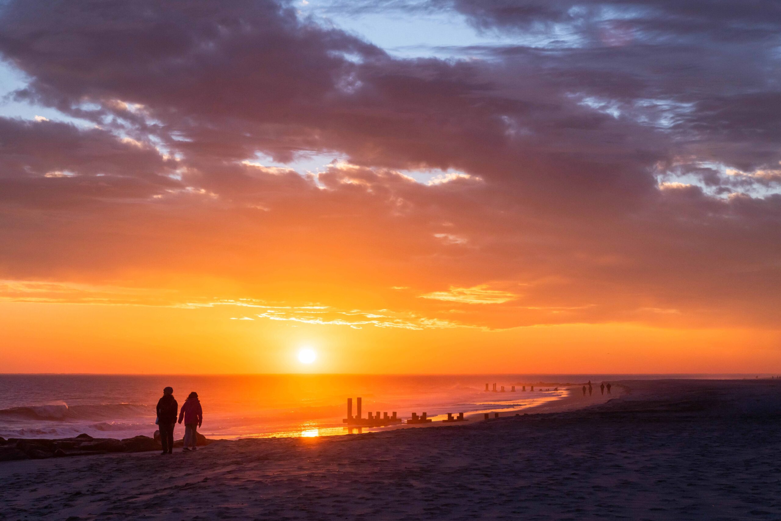 Two people walking on the beach at sunset with an orange, purple, and blue cloud in the sky