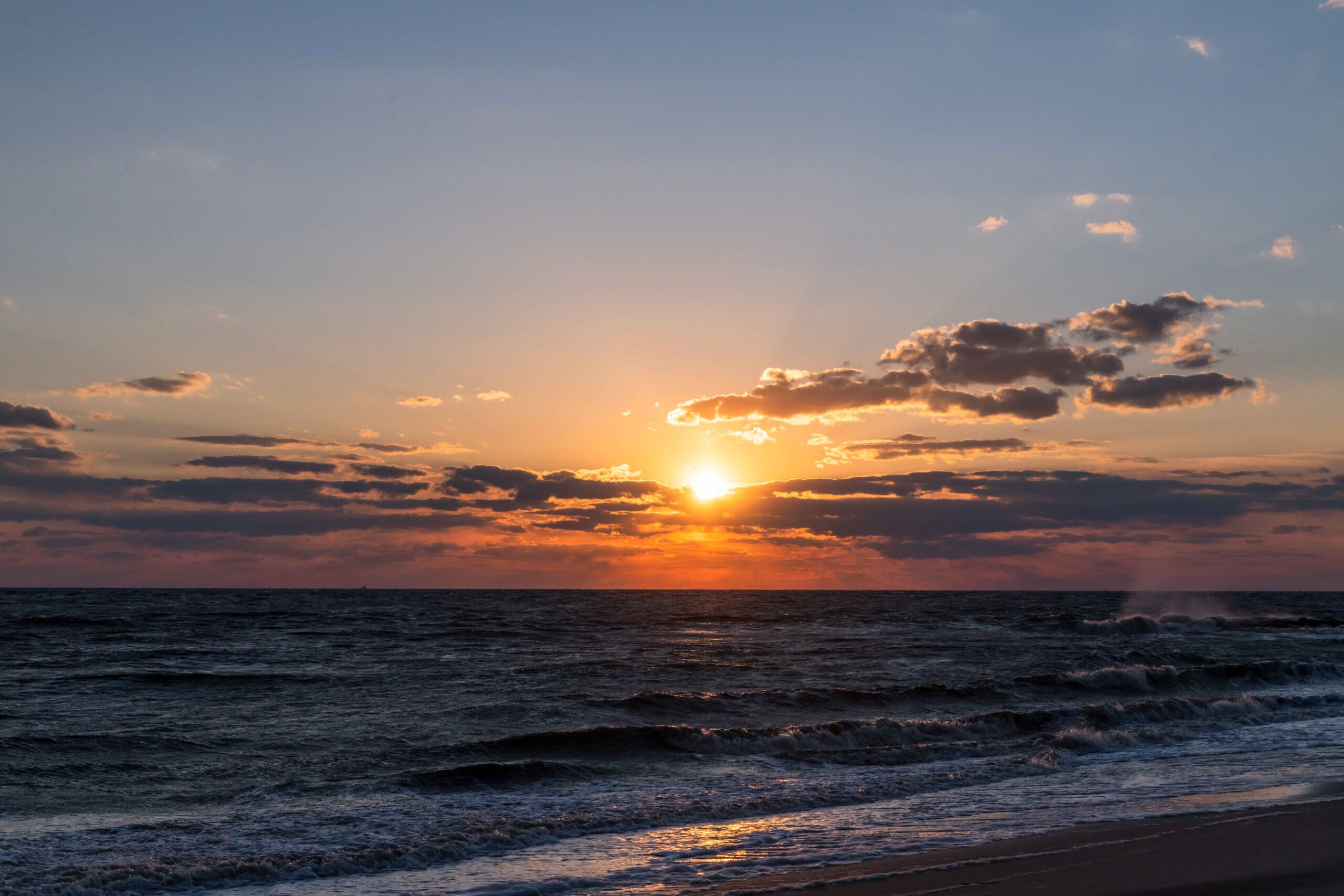 Sun setting behind some clouds at the horizon with small waves crashing in the ocean at the beach