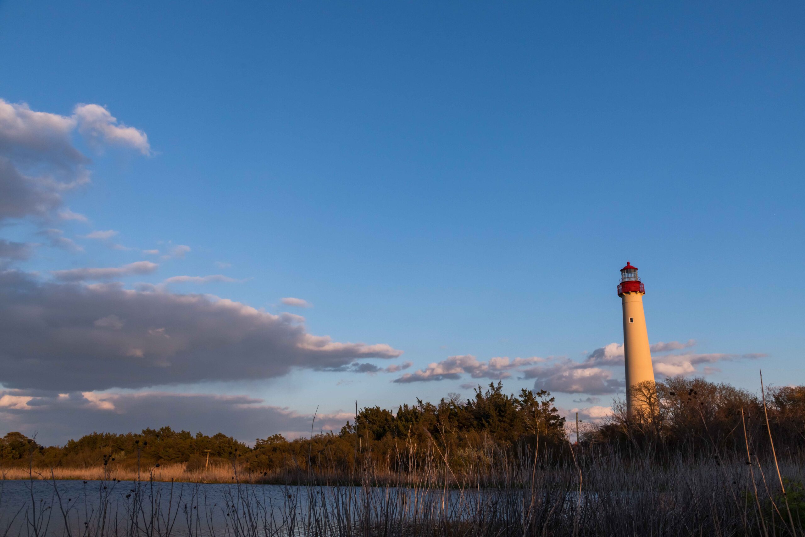 Sunlight shining on the Cape May Lighthouse at sunset with some clouds in a blue sky and a lake in the foreground