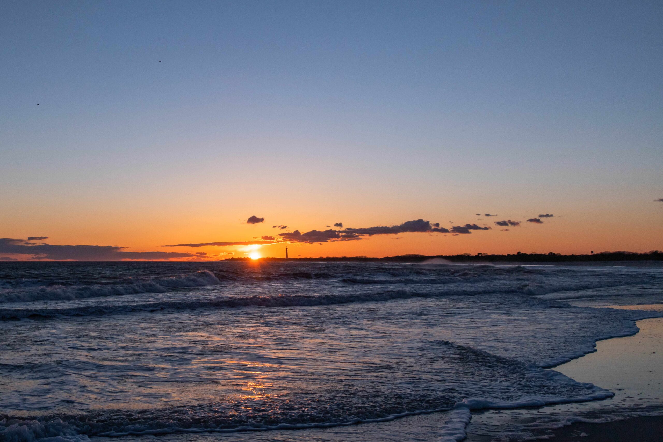 Sunset with a cool blue and orange sky with the Cape May Lighthouse in the distance, the sun reflected in the ocean, and waves rolling into the shore