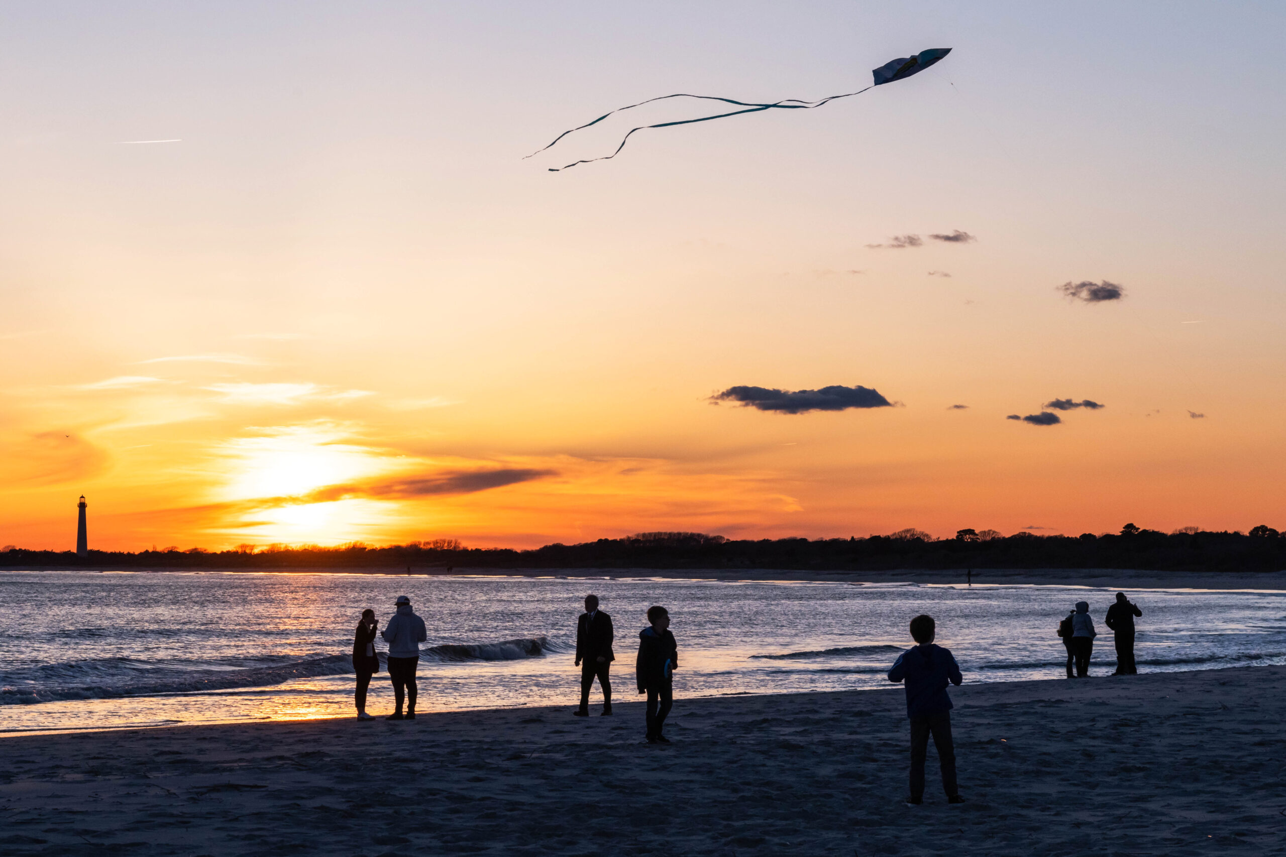 A child flying a kite at the beach at sunset with other people walking along the ocean and the Cape May Lighthouse in the distance 