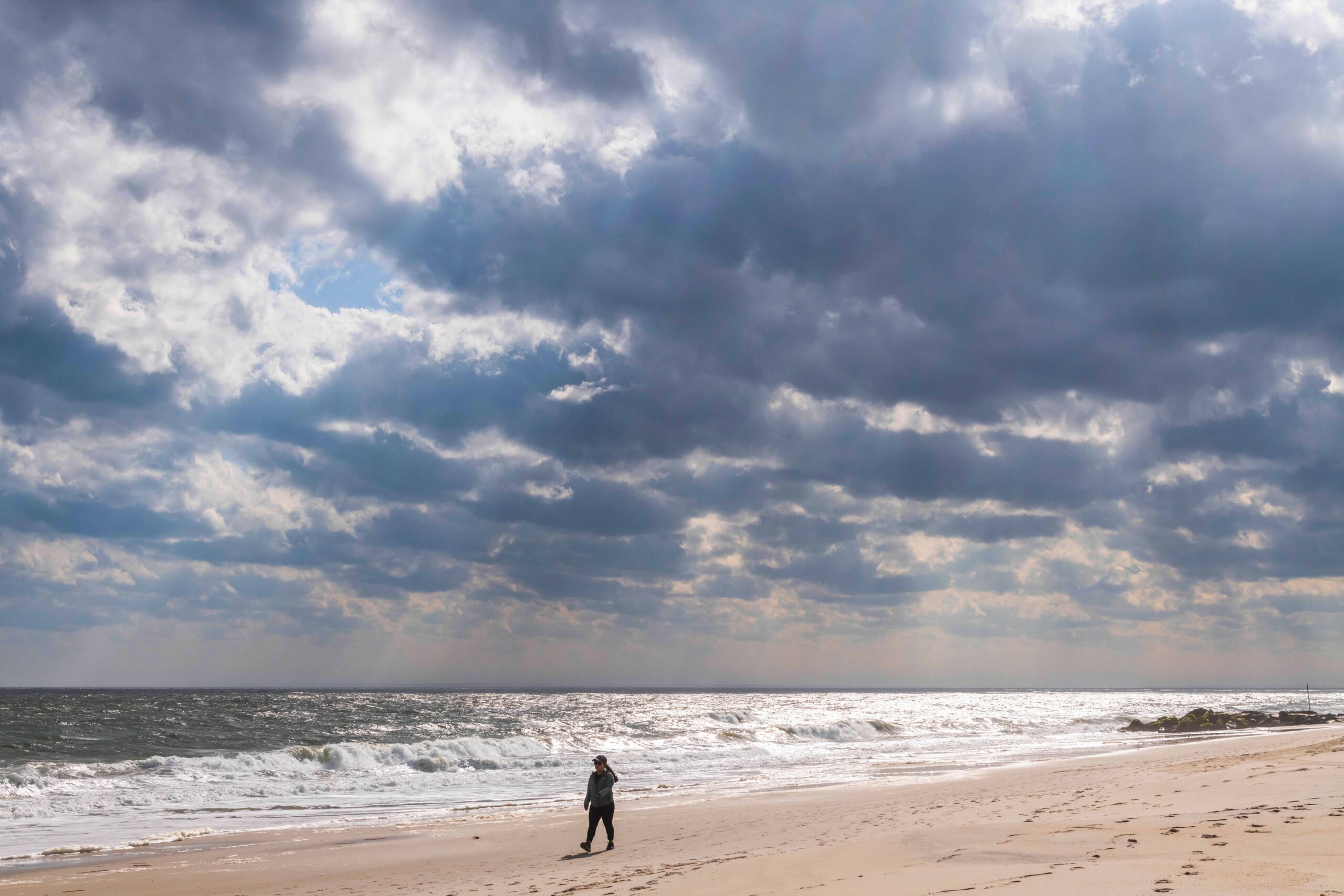 Dark puffy clouds in the sky with sun streaming through with a person walking on the beach by the ocean