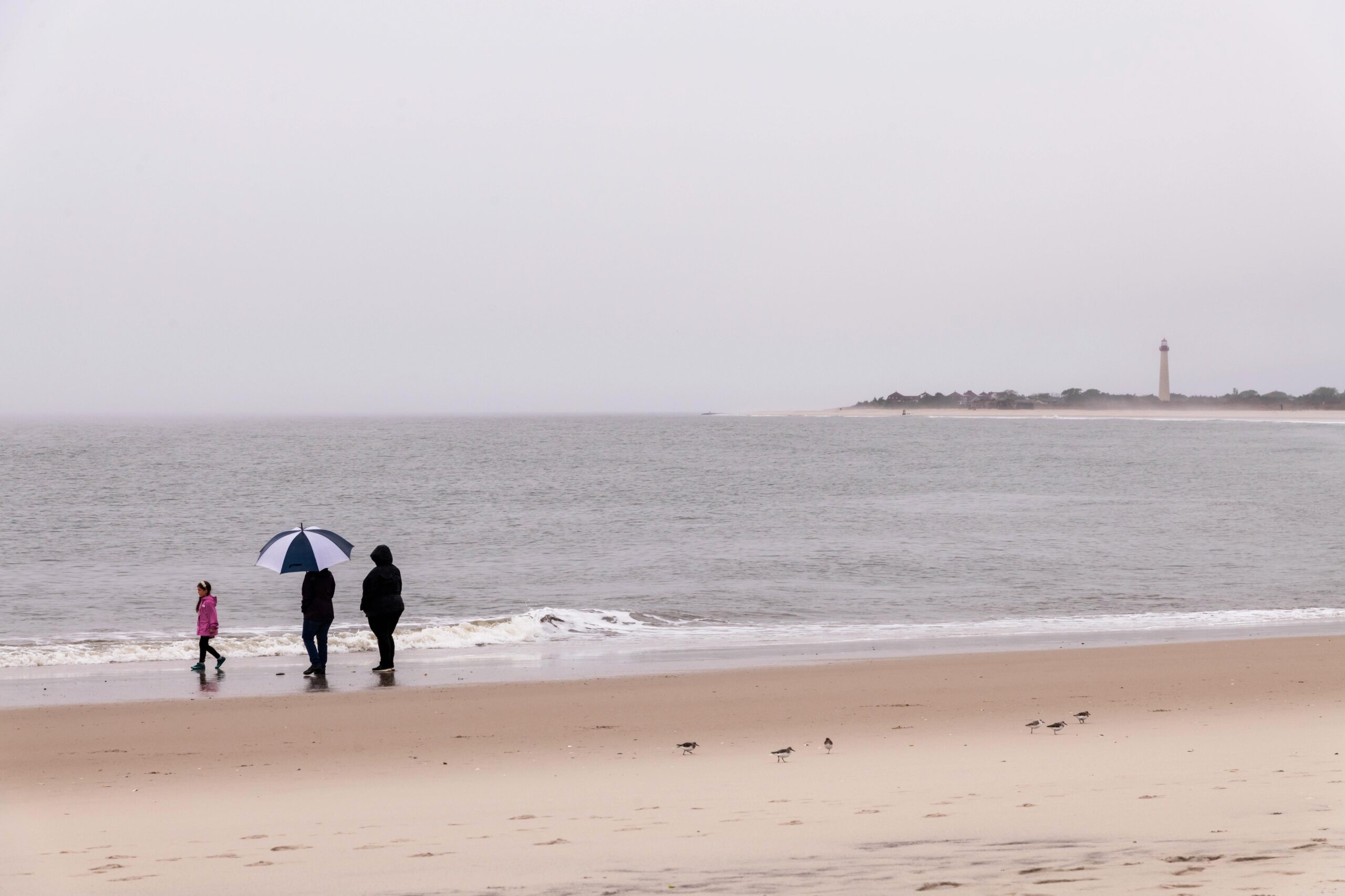 Two adults carrying an umbrella and a child walking on the beach with a cloudy gray sky and the Cape May Lighthouse in the distance