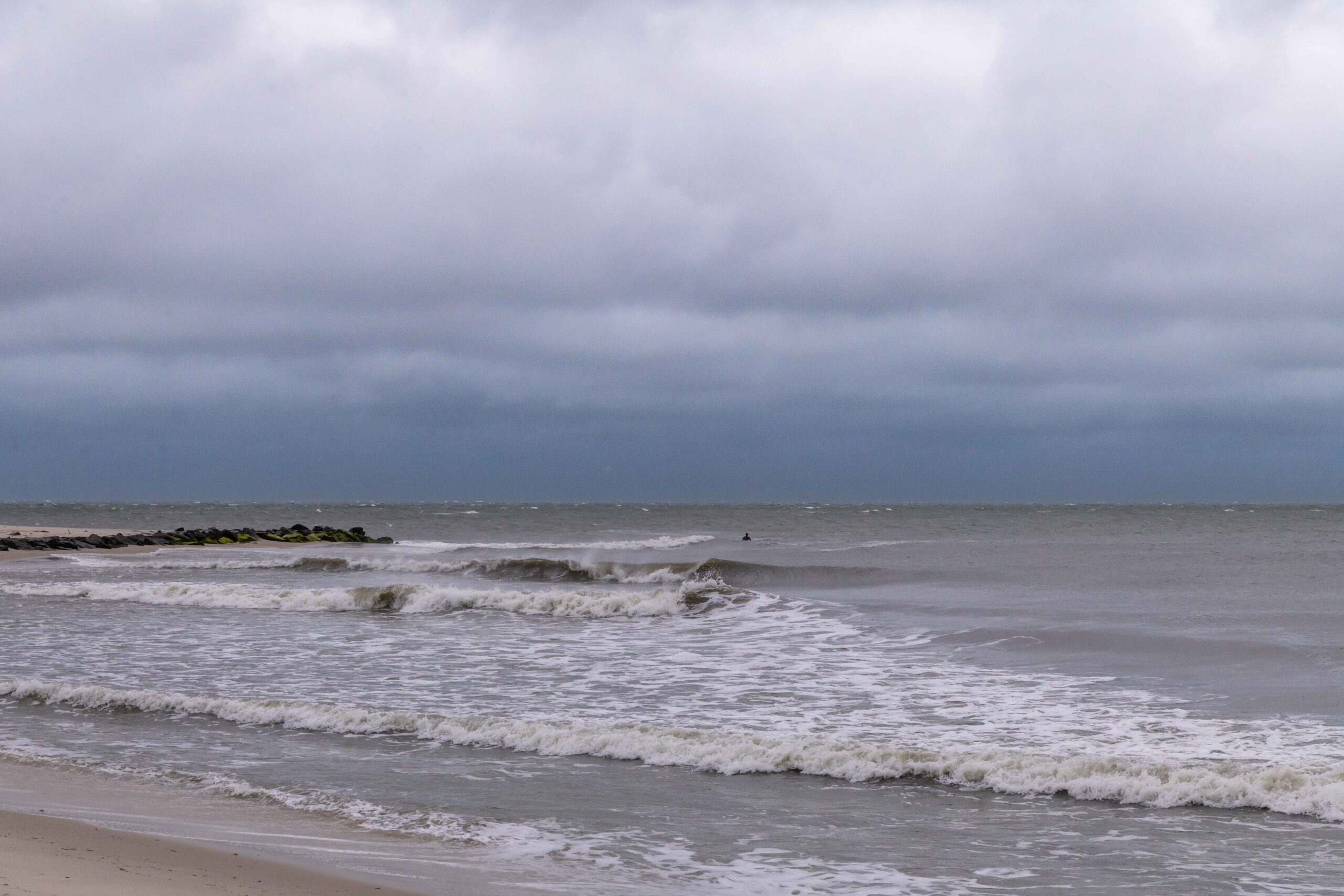 Waves crashing in the ocean on a stormy windy day with a cloudy sky and a surfer out in the distance