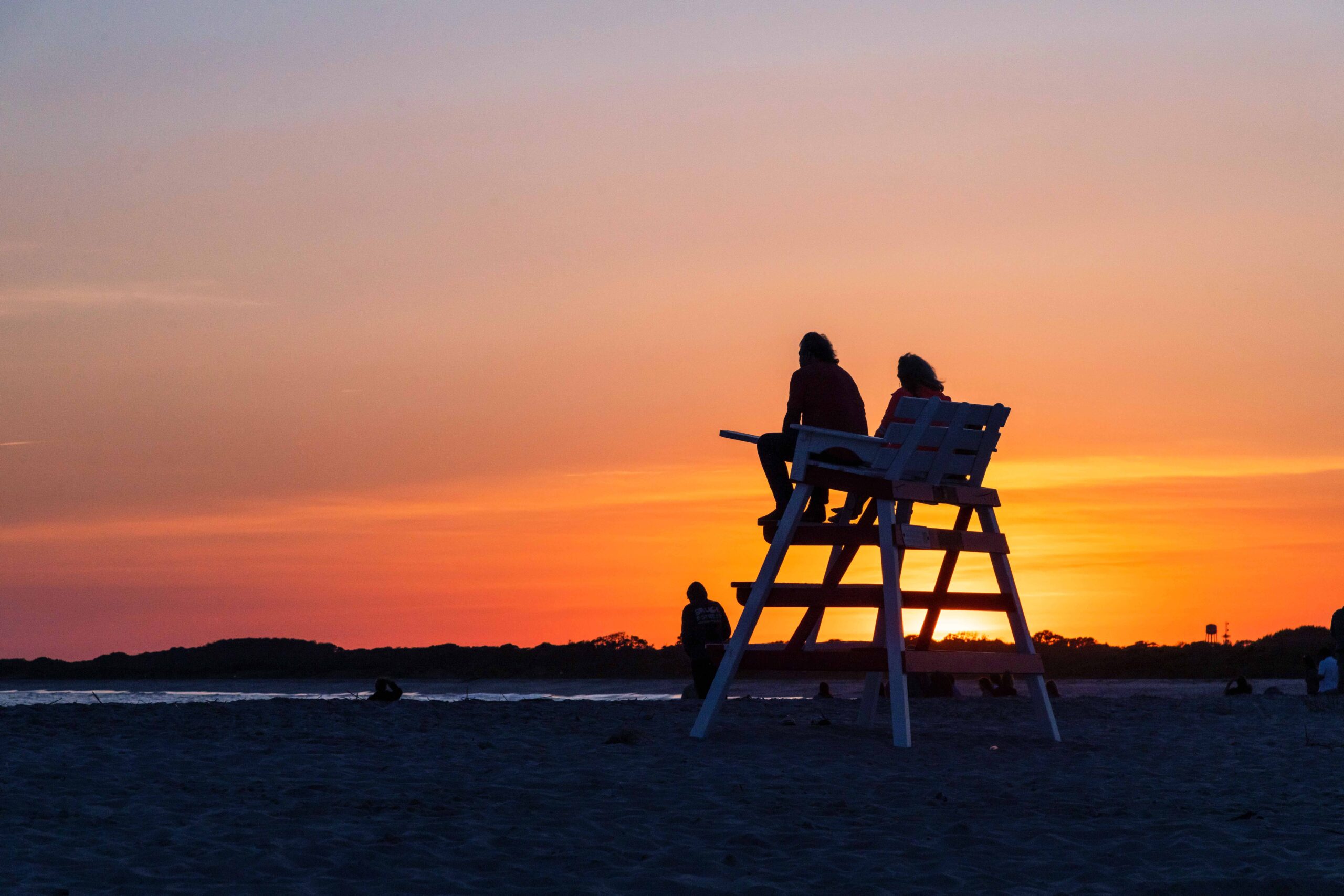 Two people sitting on a lifeguard stand silhouetted by sunset at the beach