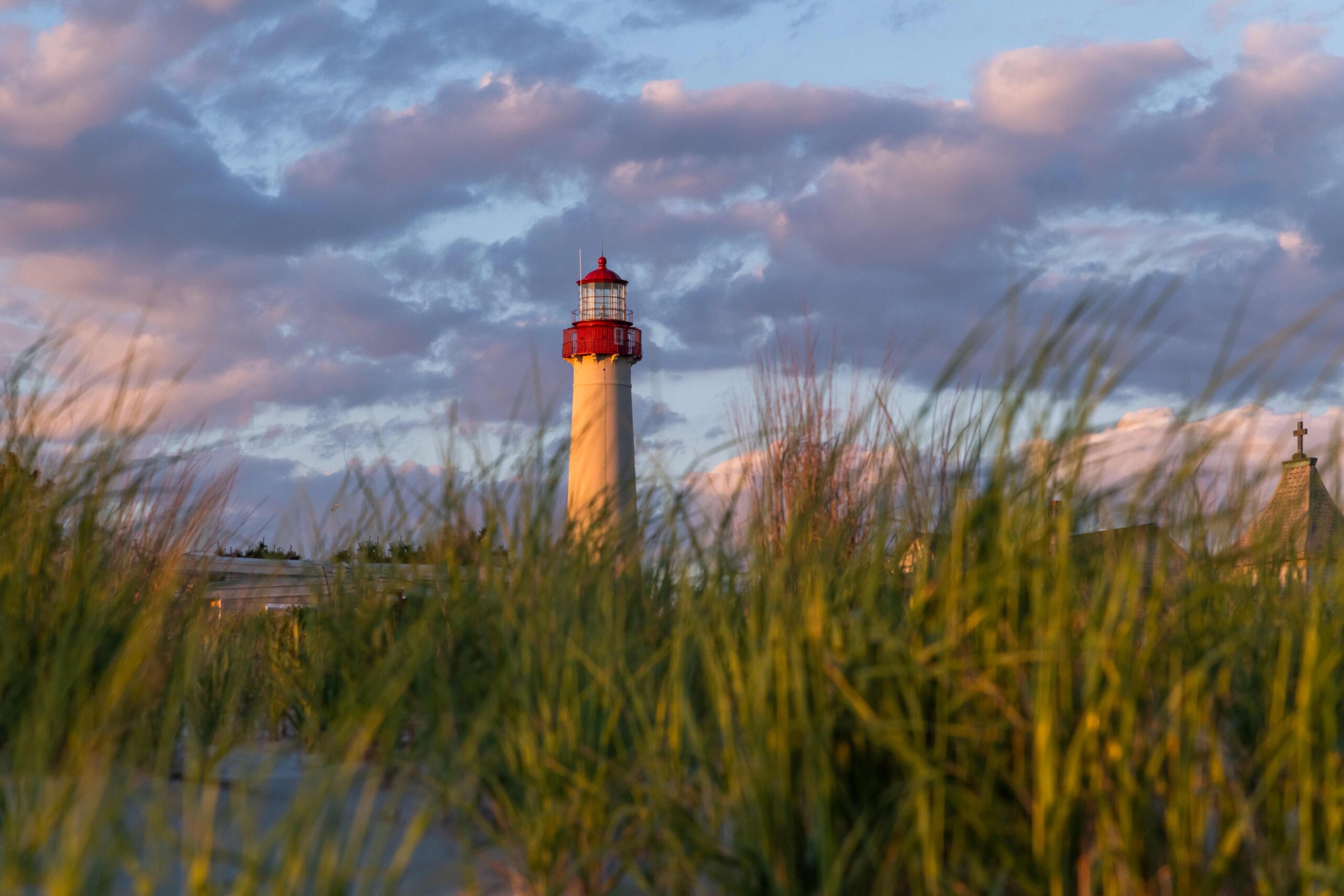 Sunset light shining on the Cape May lighthouse with purple and pink clouds in the sky and green beach dunes in the foreground
